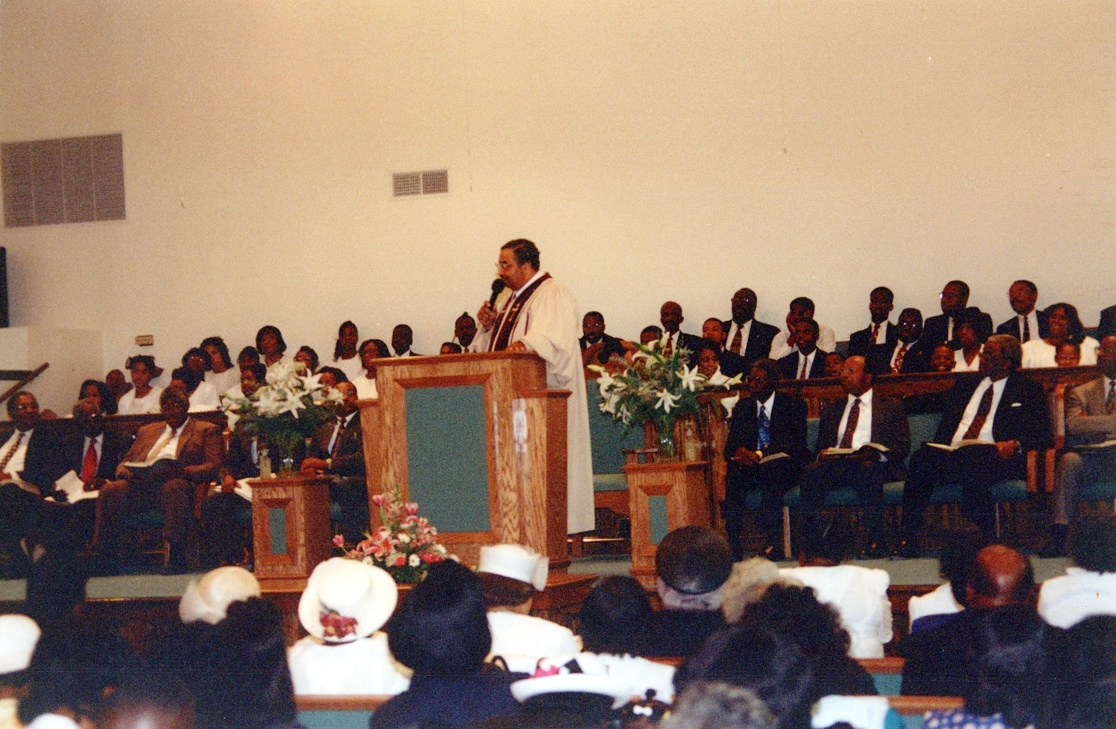 A Black pastor in a white robe stands at a wooden pulpit, speaking to a large congregation seated in pews. The photograph has a grainy, vintage quality.