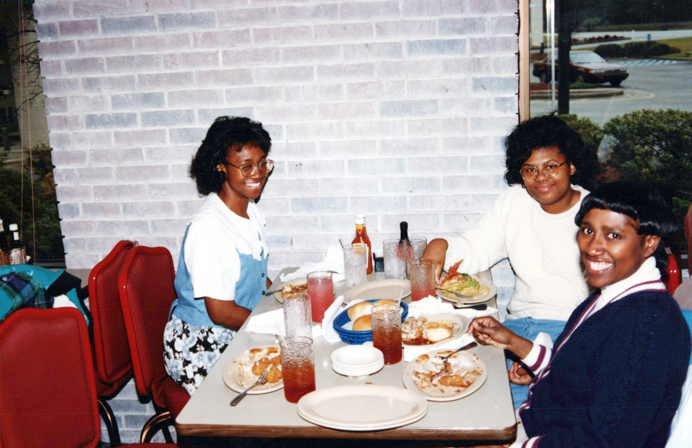 A vintage photo of three smiling African American women sitting at a table in a restaurant, enjoying a meal together against a light brick wall.