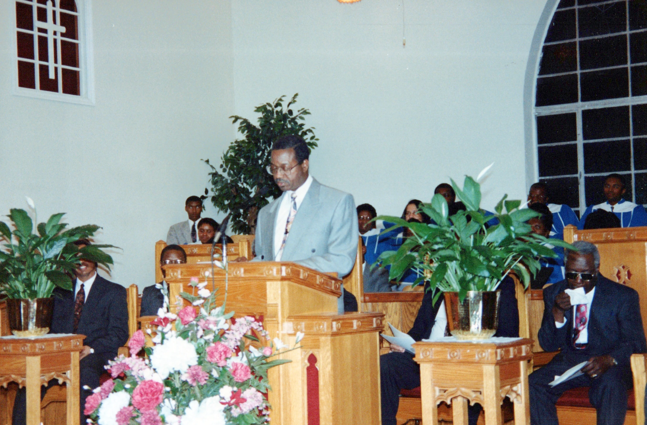An older Black man in a light grey suit and glasses stands at a wooden pulpit, speaking to a congregation during a church service.