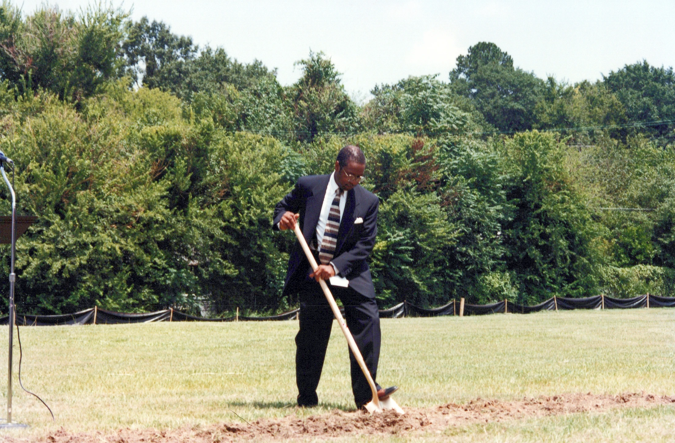 An African American man in a dark suit and tie uses a shovel to dig into the dirt in a grassy field, marking the start of a new project.