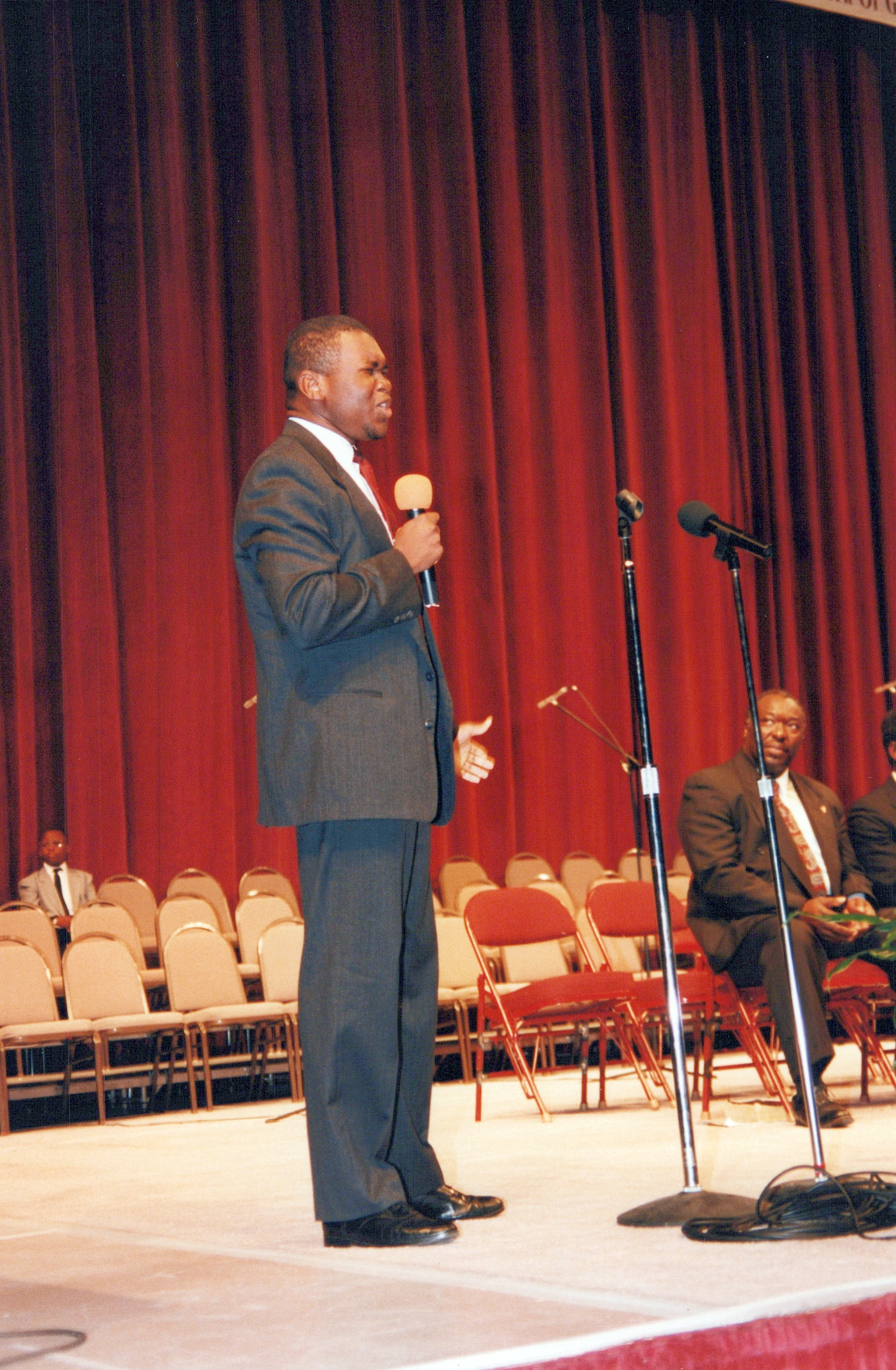 A Black man in a dark suit stands on a stage in front of a red curtain. He is holding a microphone and speaking passionately. Others are seated behind him.