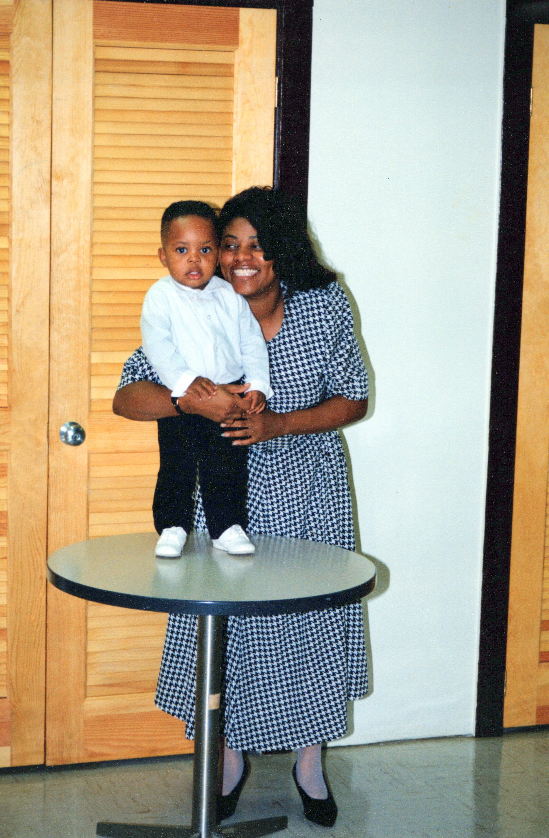 A smiling Black woman in a houndstooth dress holds a young boy who is standing on a round table. The boy wears a white shirt, black pants, and white shoes.