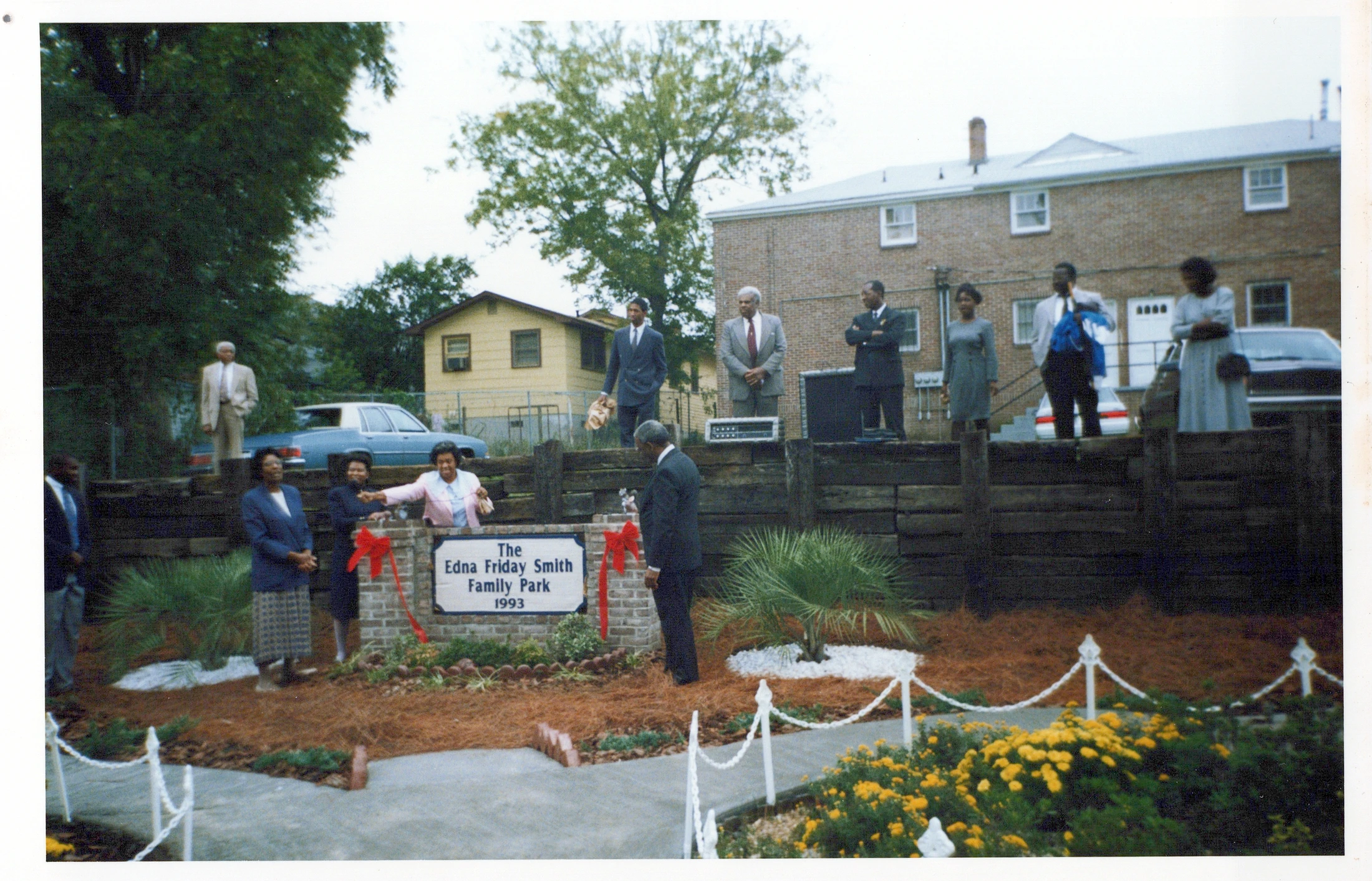 A group of formally dressed African American people attend the 1993 dedication of The Edna Friday Smith Family Park, gathered around a brick sign with red bows.