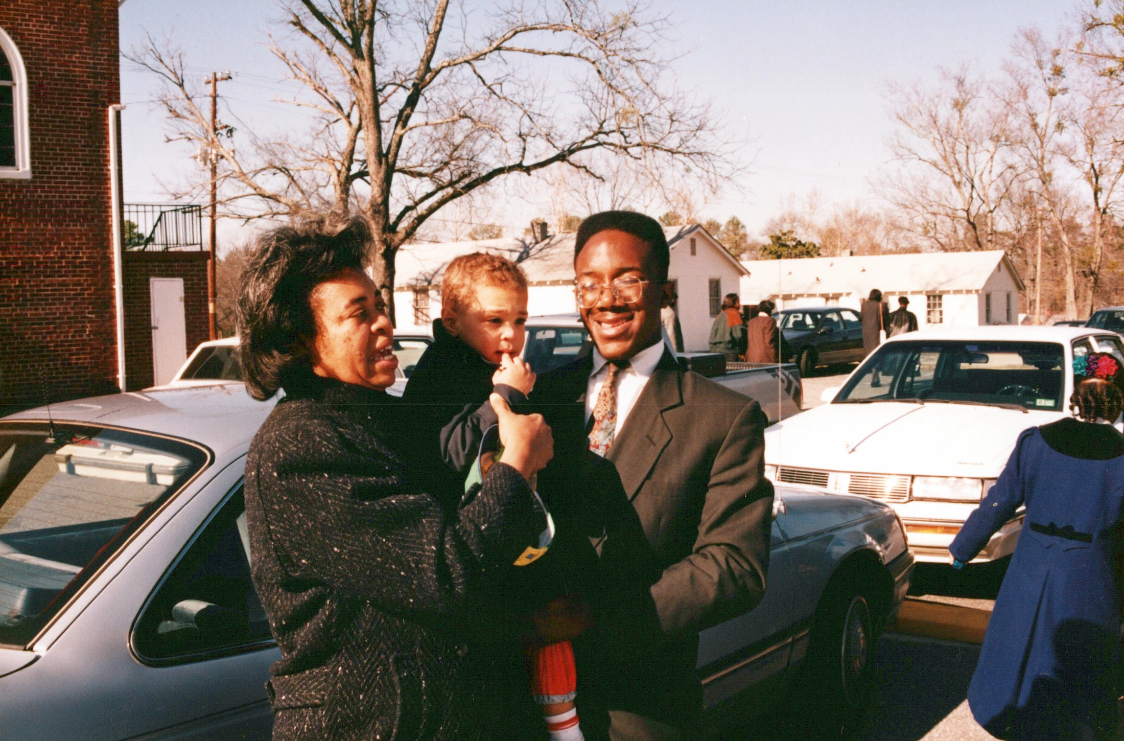 A vintage photo of a Black family, a woman and a man in a suit, smiling as they hold a young child together in a parking lot on a sunny day.