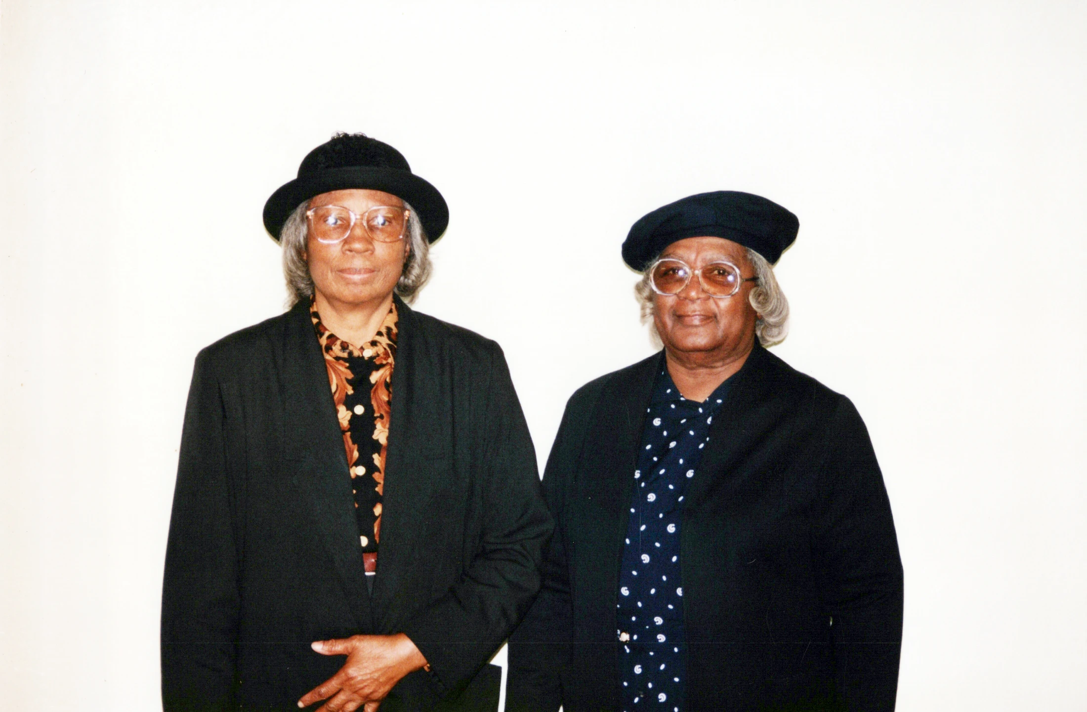 A waist-up portrait of two elderly African American women standing together. Both wear dark jackets, glasses, and black hats, smiling gently at the camera.