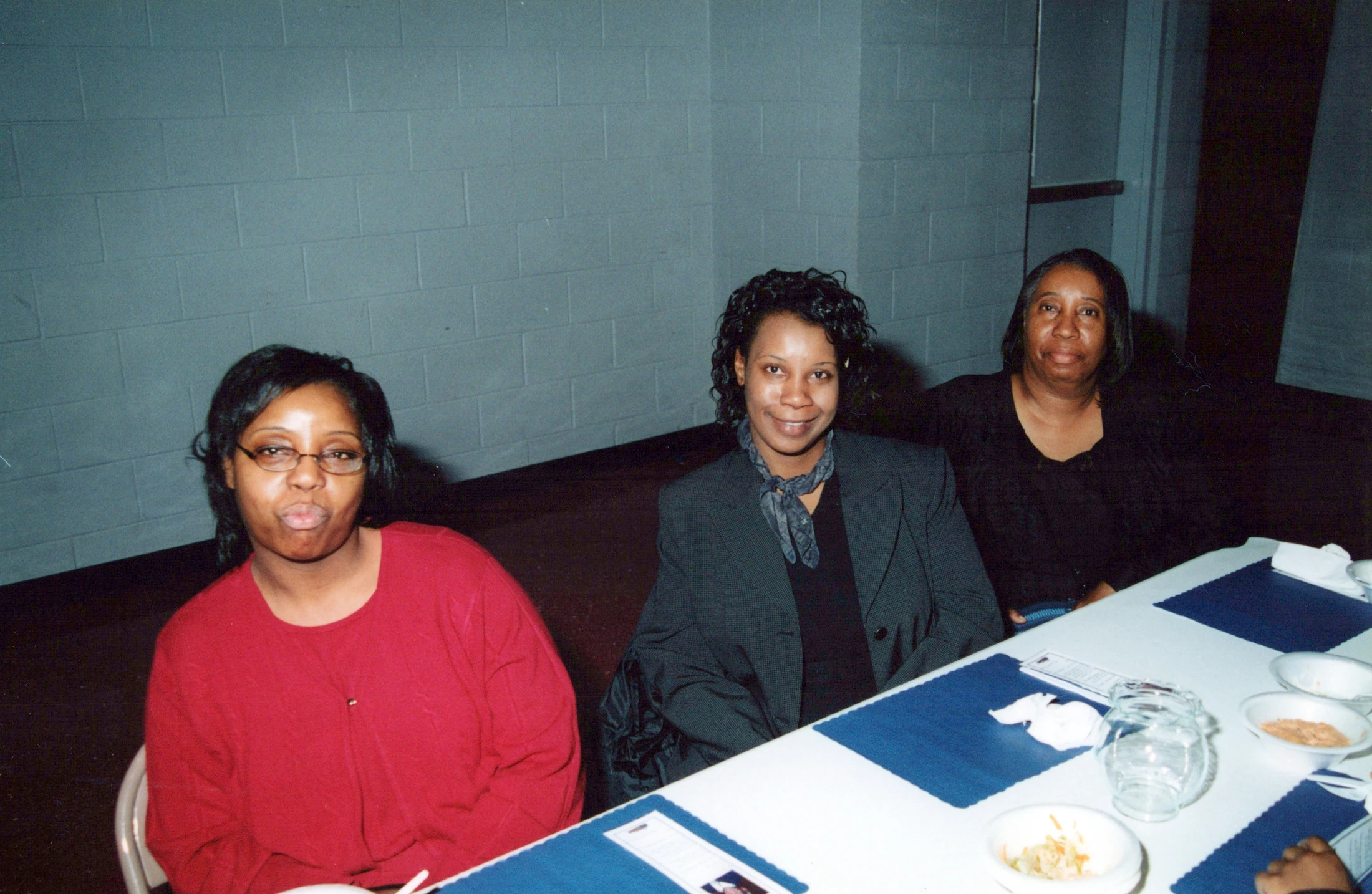 Three Black women sit side-by-side at a dining table with blue placemats, looking at the camera. The background is a painted grey cinder block wall.