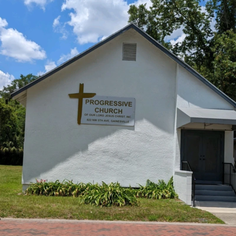A small white church building with a gabled roof against a blue sky. A sign identifies it as the Progressive Church, with a golden cross above the sign.