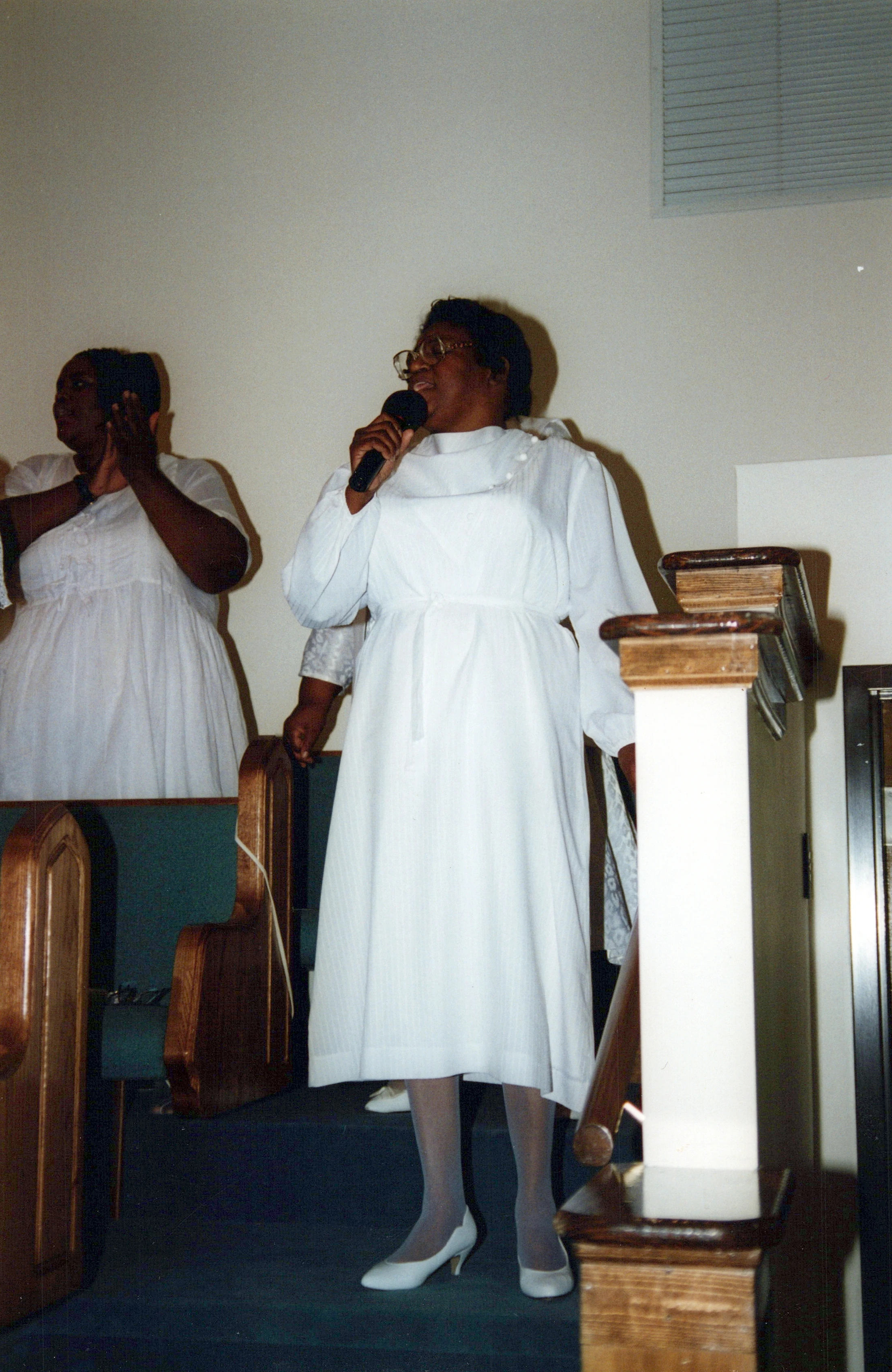A Black woman in a white dress and glasses sings into a microphone in a church. Another woman in a white dress claps behind her near wooden pews.