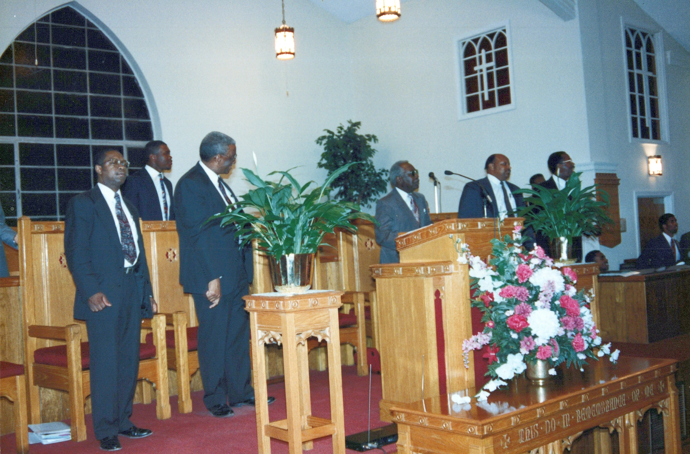 A group of Black men in dark suits stand at the front of a church sanctuary near a wooden pulpit, communion table, and floral arrangements.