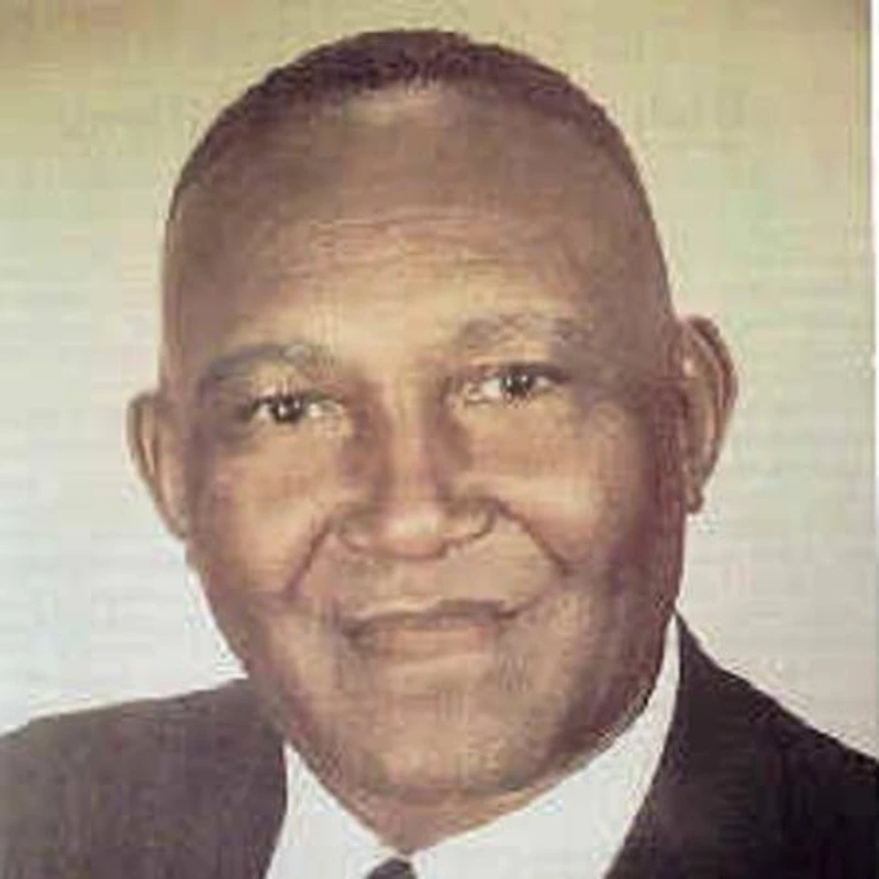 A warm, color headshot of an older Black man with a shaved head. He is smiling slightly while looking at the camera, wearing a dark suit and white shirt.