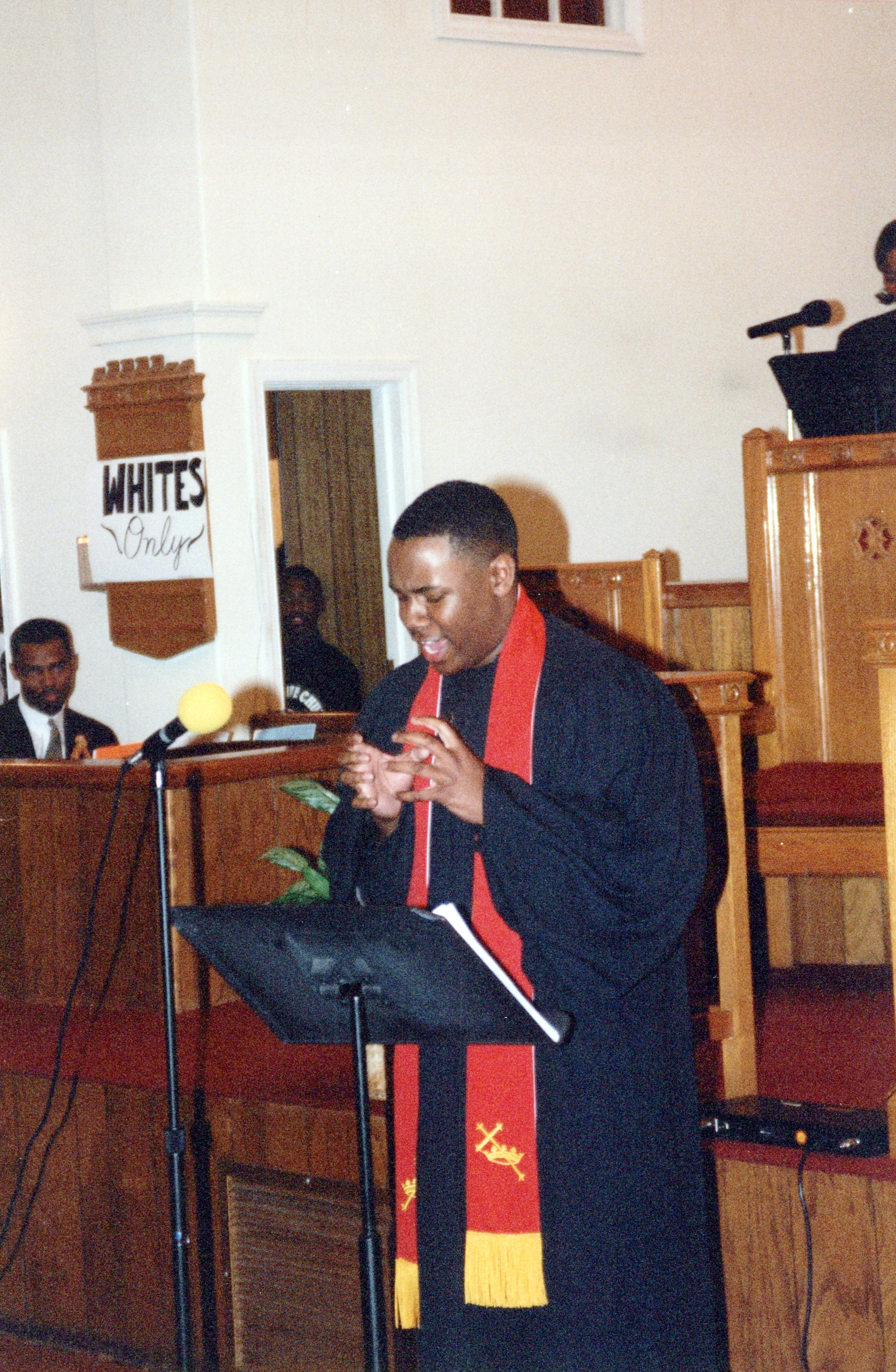 A Black minister in a robe preaches from a podium in a church. A sign that reads 'WHITES Only' is visible in the background, likely part of a performance.
