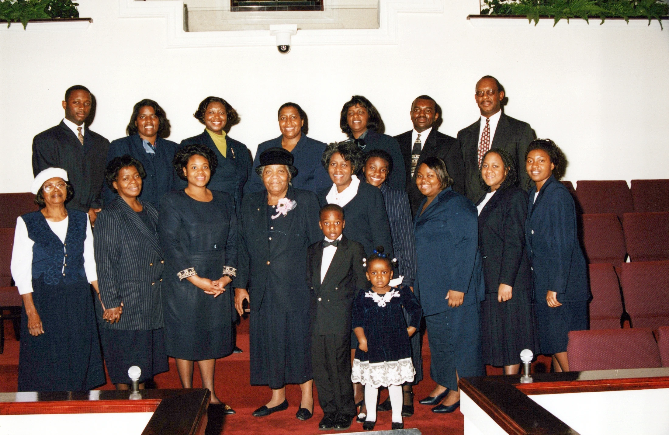 A large, multi-generational group of African American church members dressed in formal attire poses for a group photograph in a church sanctuary.