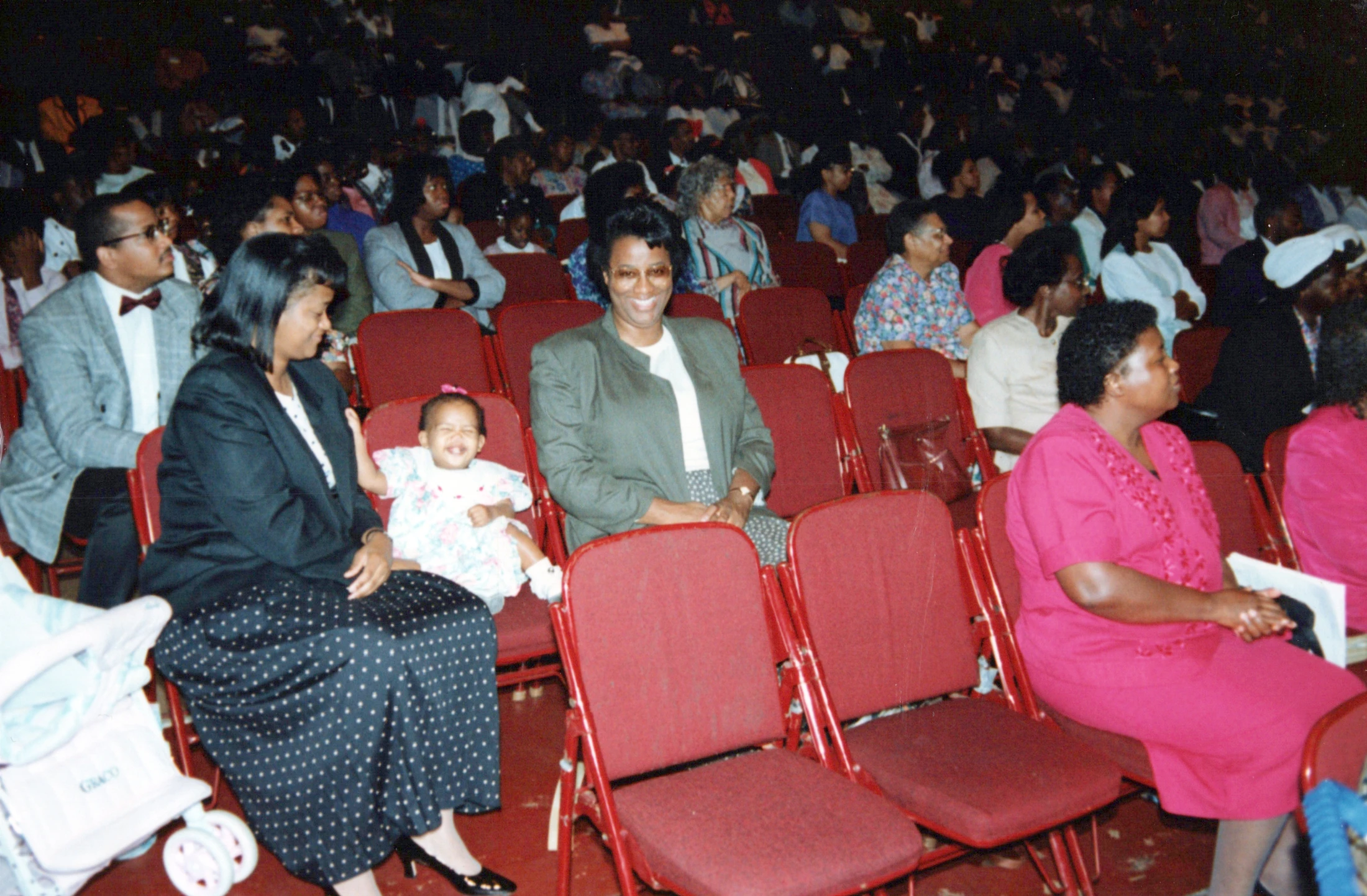 A vintage photograph of a large audience of Black people seated in red chairs. In the foreground, a baby girl smiles happily on a woman's lap.