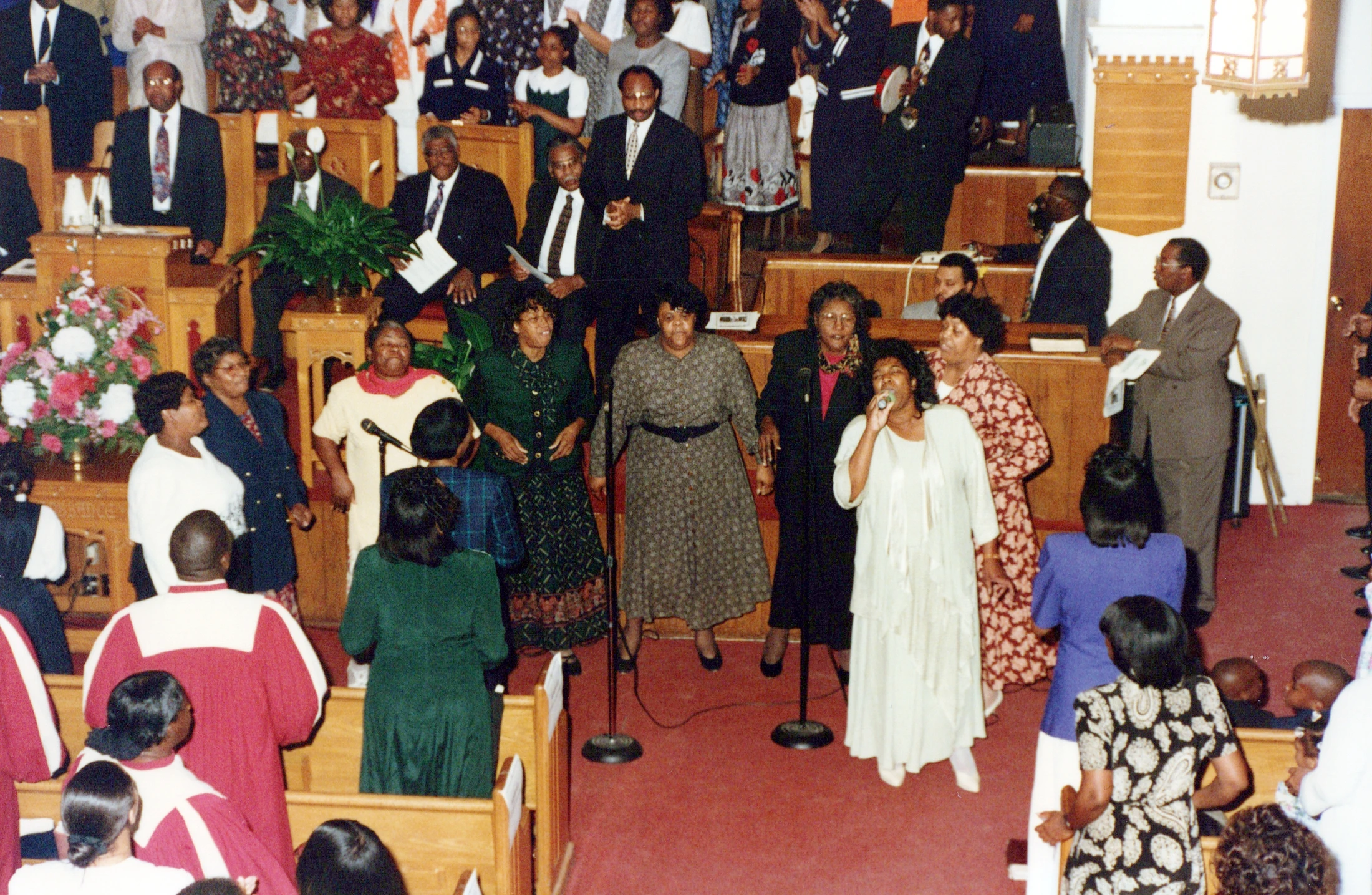 A Black church choir performs during a service. A woman in a white dress sings lead into a microphone while other members sing alongside her.