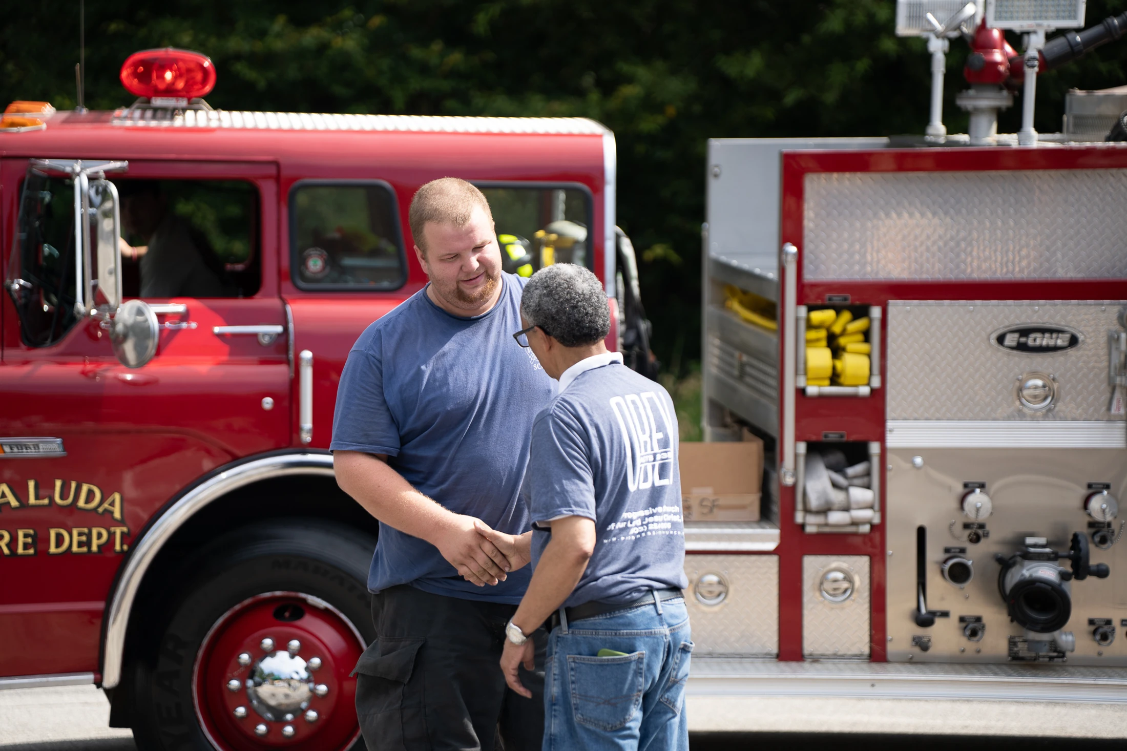 A firefighter and another man shake hands while standing between two red fire trucks from the Saluda Fire Department on a sunny day.
