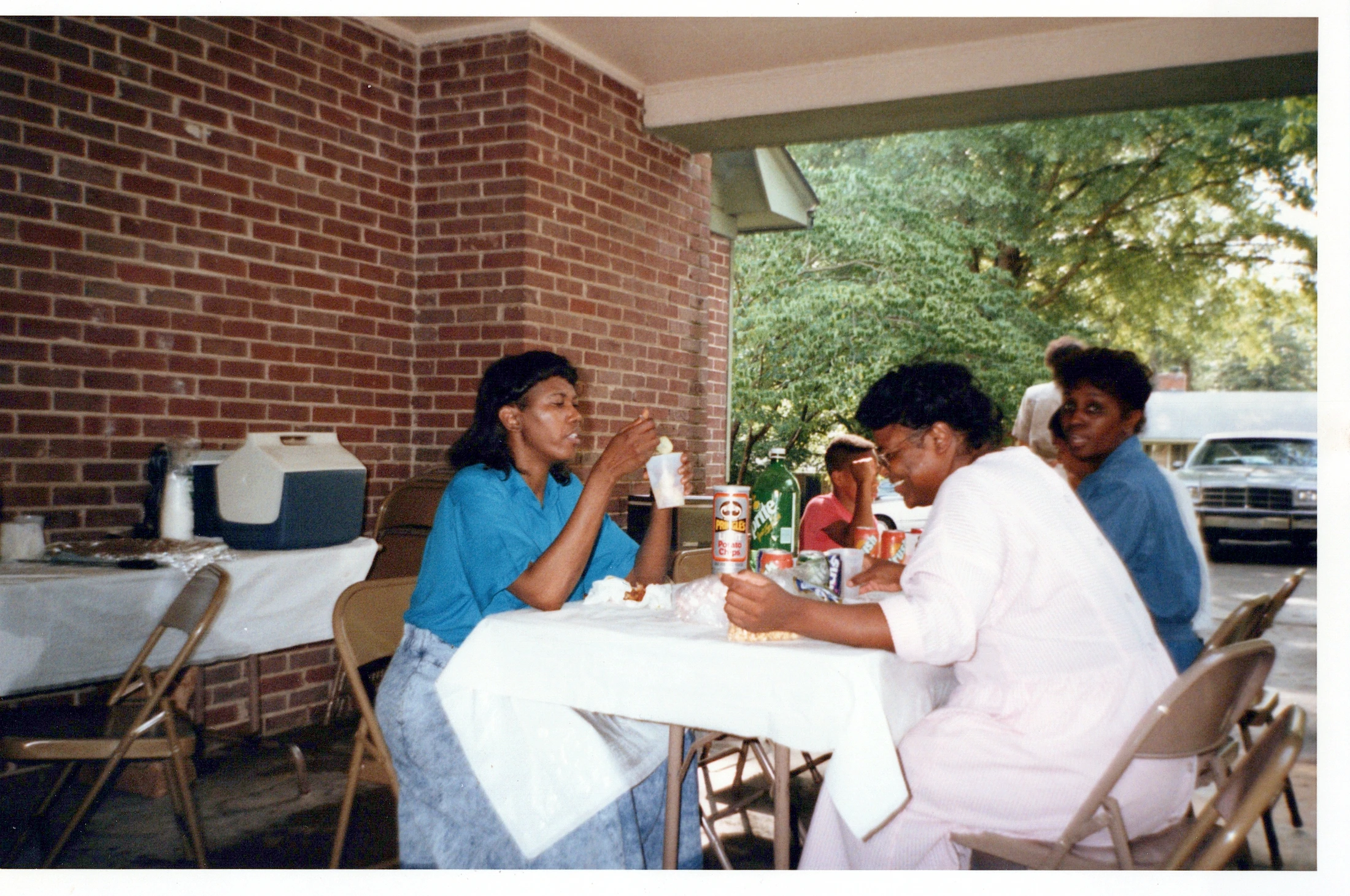 A vintage photograph of several Black women sitting at a white-clothed table on a patio, eating and talking during a sunny day gathering.