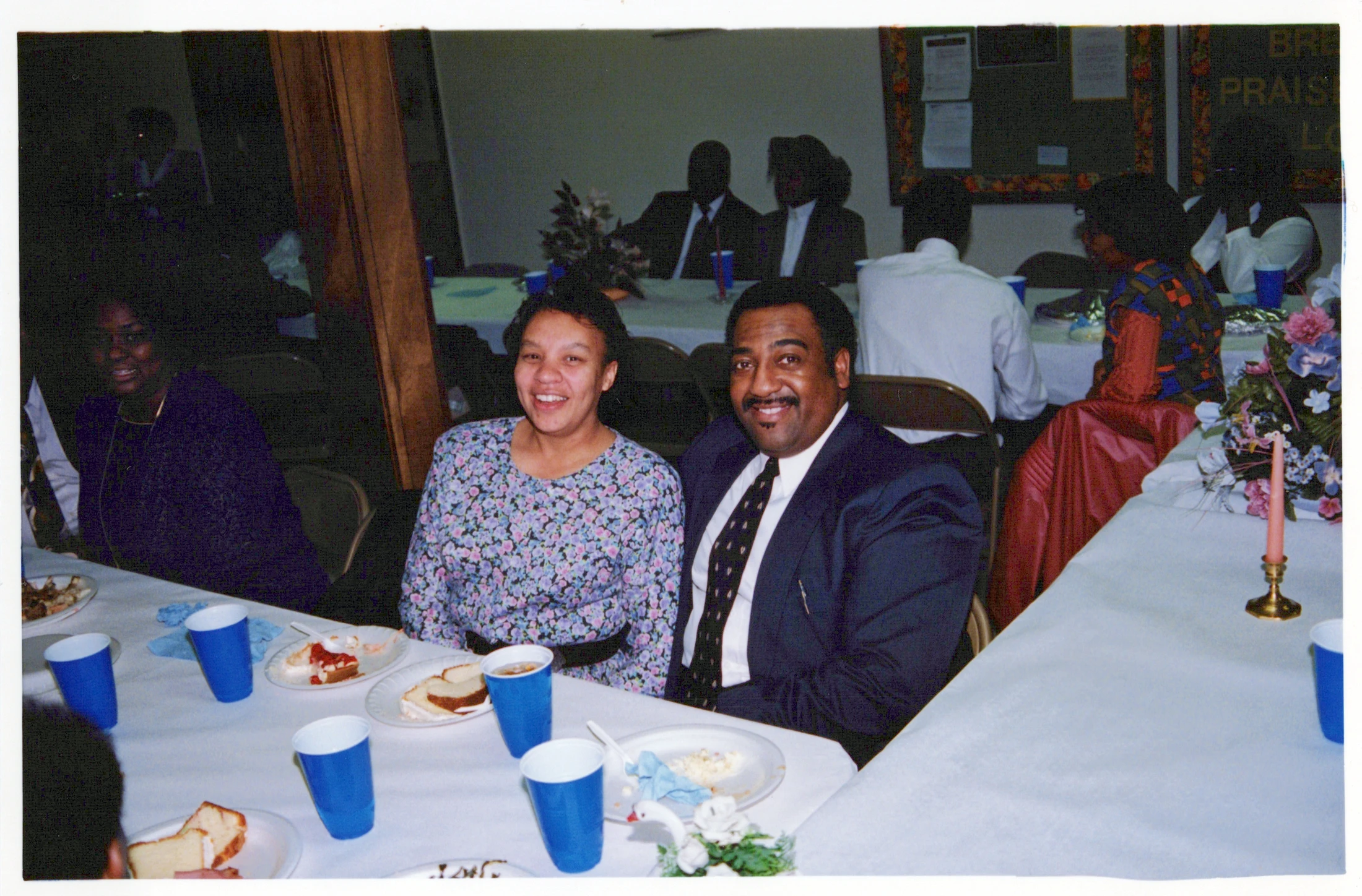 An African American couple smiles for the camera while seated at a long, white-clothed table during a banquet with other guests in the background.