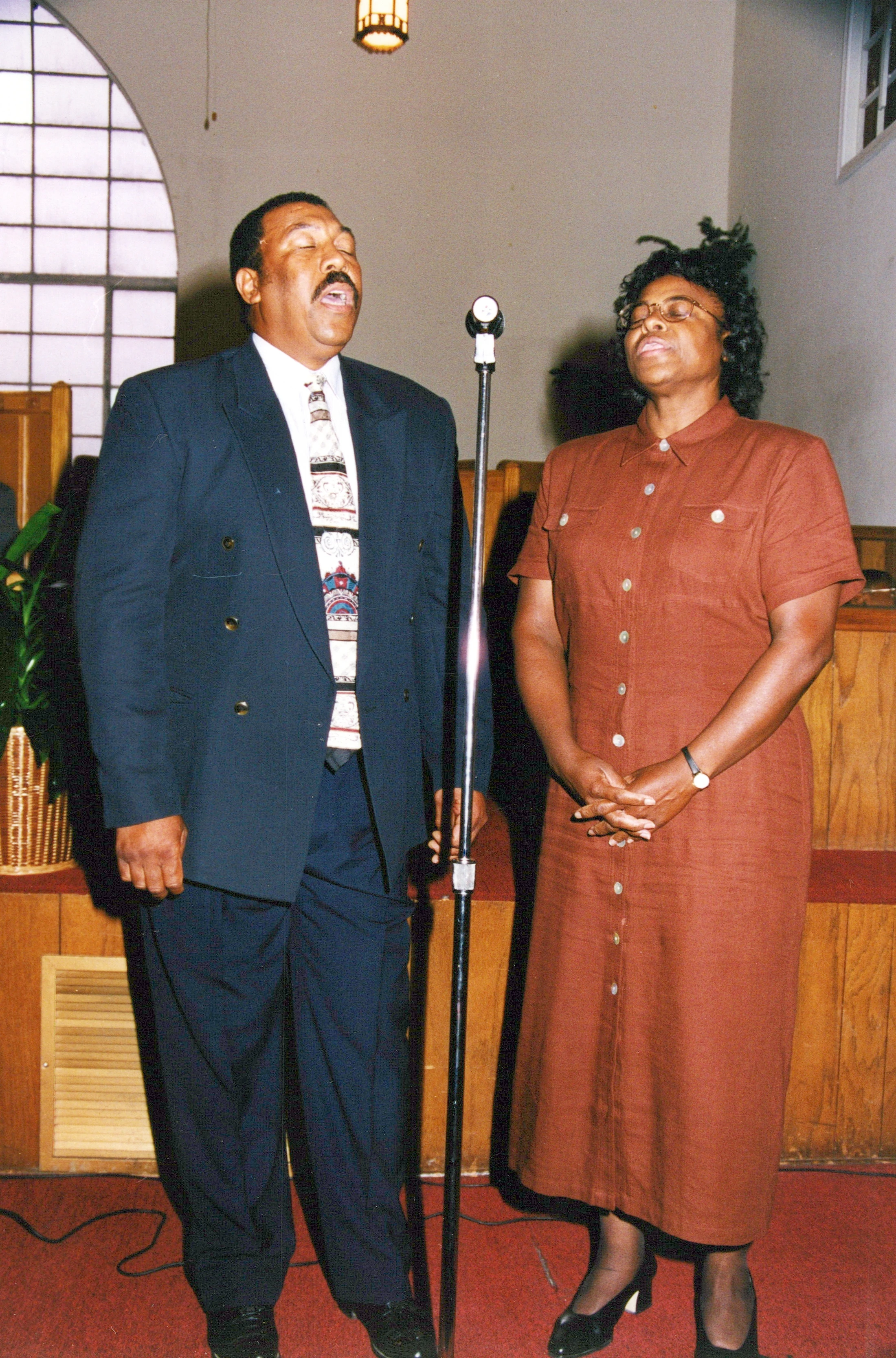 An African American man and woman sing with their eyes closed at a microphone stand inside a church with wood paneling and red carpet.
