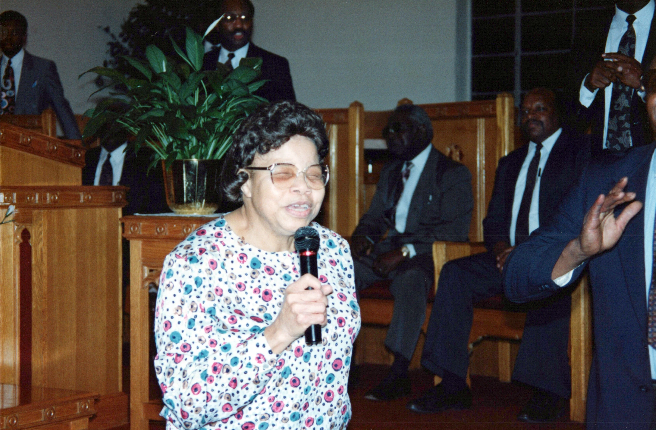 A Black woman in a colorful blouse and glasses speaks into a microphone in a church. In the background, several men in suits are seated on wooden pews.