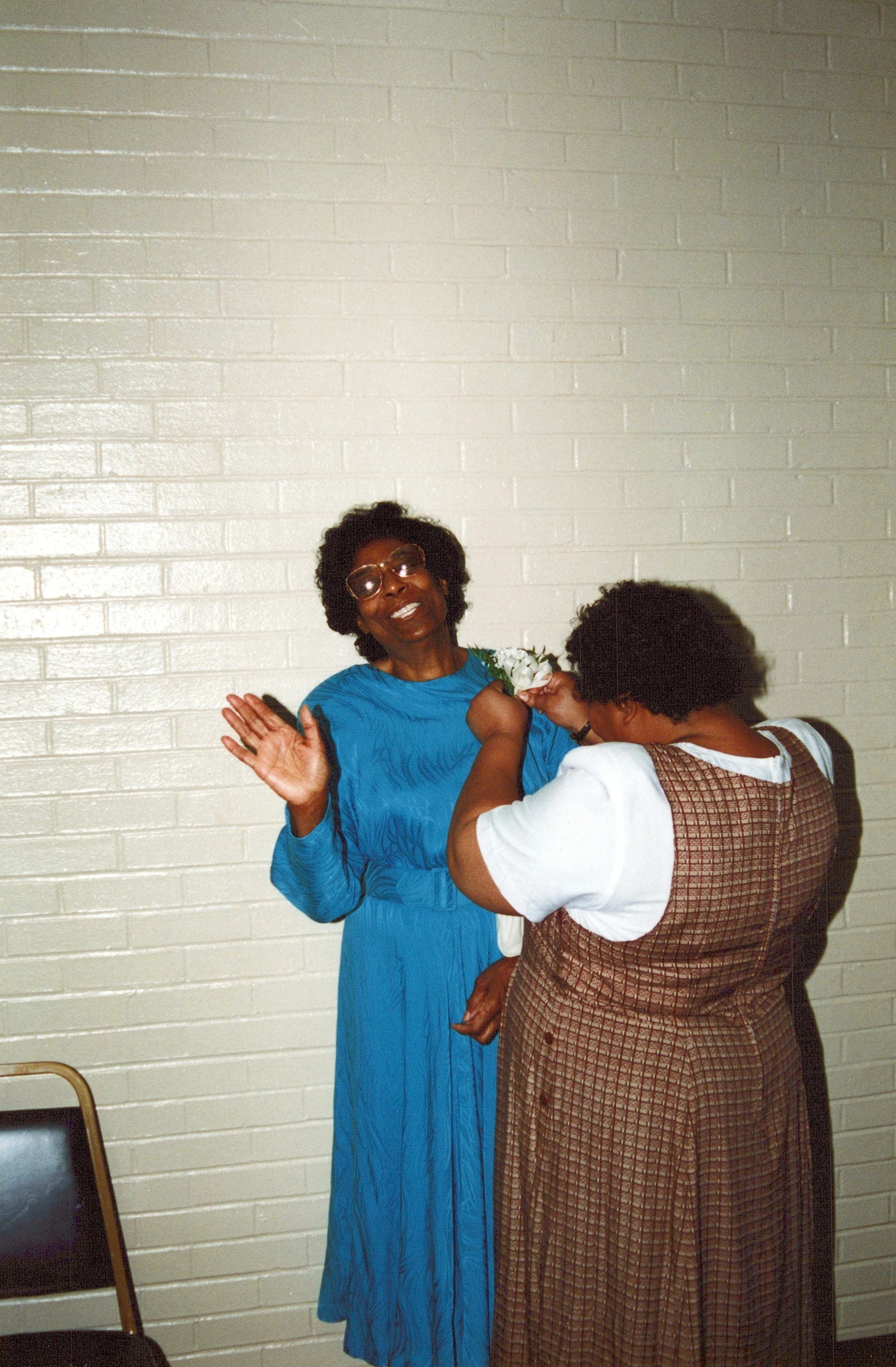 A vintage photo of two Black women against a white brick wall. One woman in a blue dress smiles as another pins a white flower corsage on her shoulder.