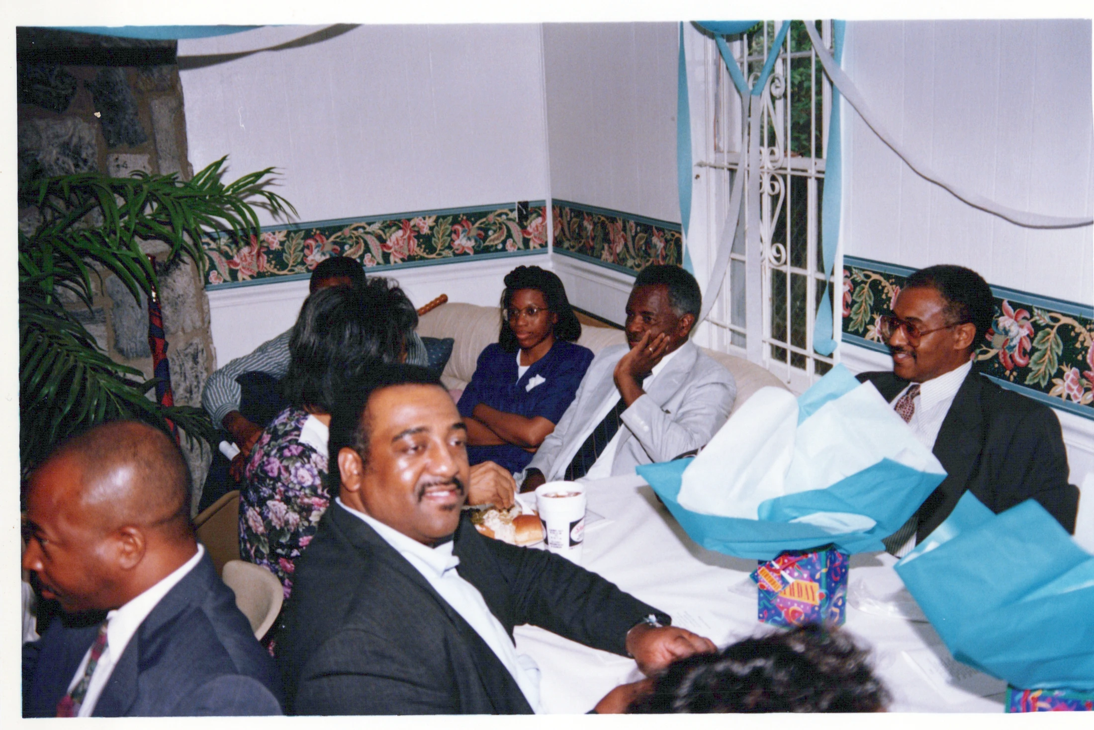 A candid photo of a group of Black adults sitting around a table at an indoor party. Gifts are on the table, and they are dressed for a celebration.