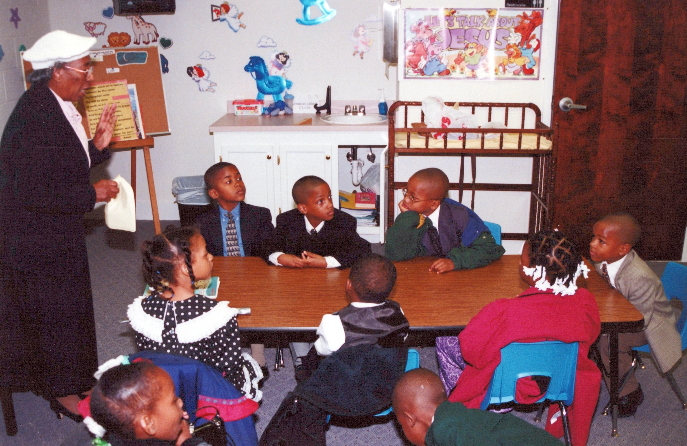 An older Black woman in a white hat teaches a group of young Black children who are sitting attentively around a wooden table in a classroom.