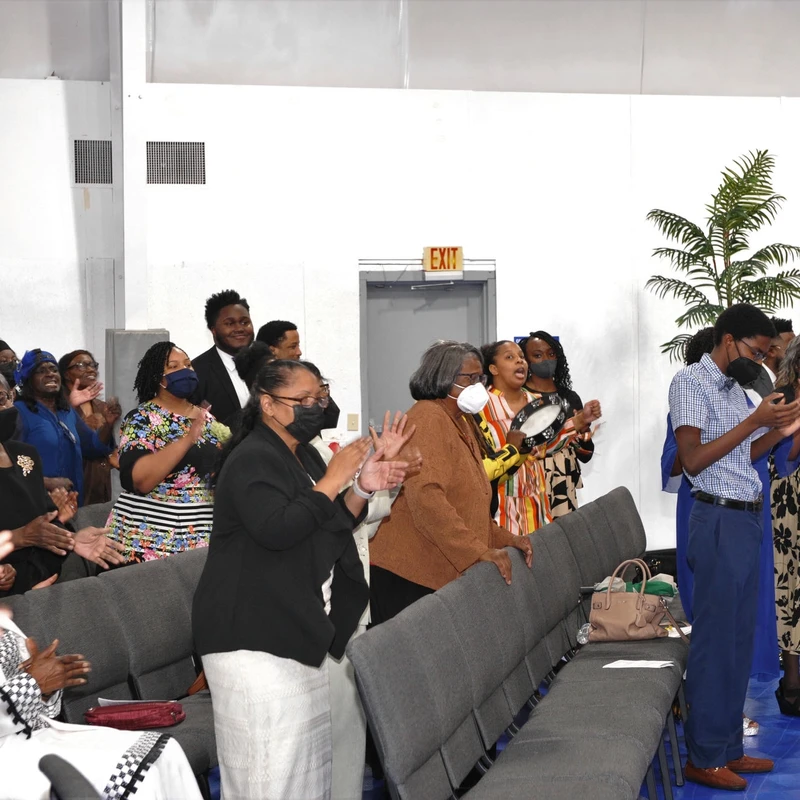A group of Black church members, many wearing face masks, stand and clap their hands joyfully during a service in a large room with grey chairs.