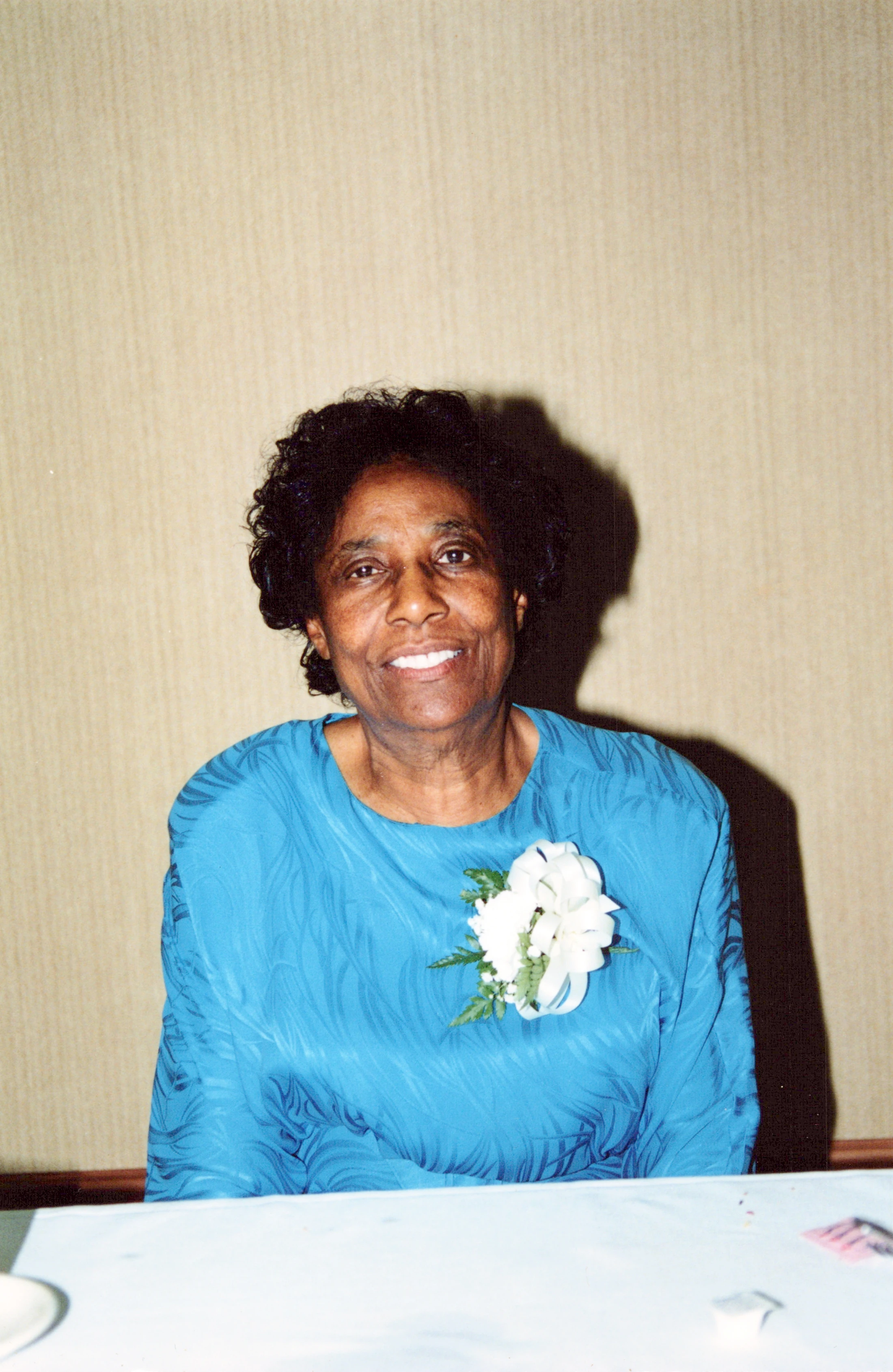A smiling older Black woman with short, dark curly hair, wearing a vibrant blue top with a white flower corsage, sits at a table against a neutral wall.