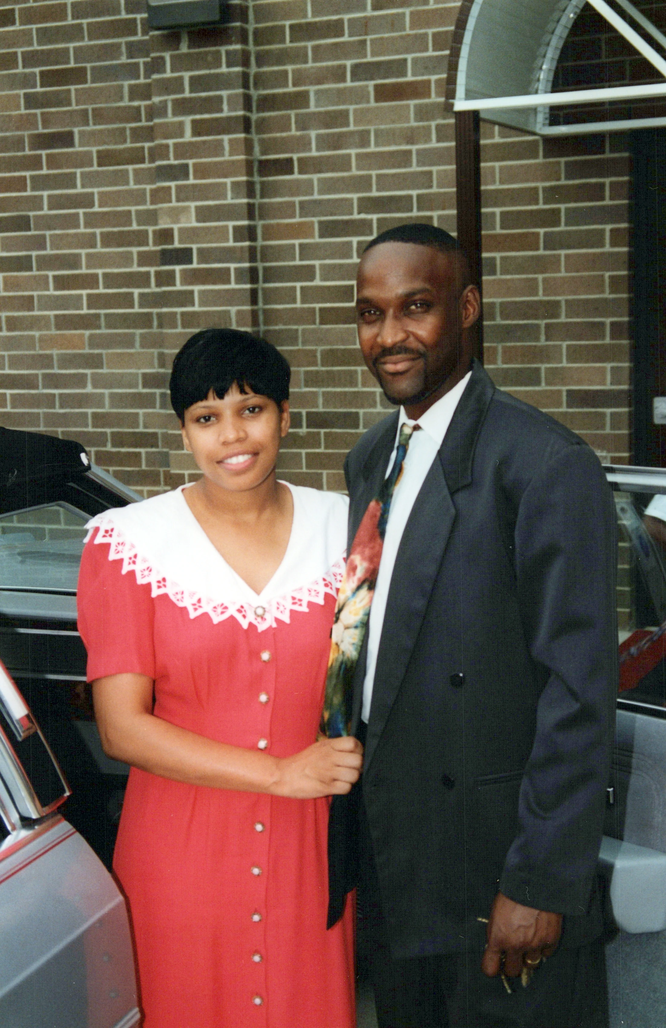 A smiling Black man in a dark suit and a Black woman in a red dress with a white lace collar pose together outdoors in front of a brick wall.