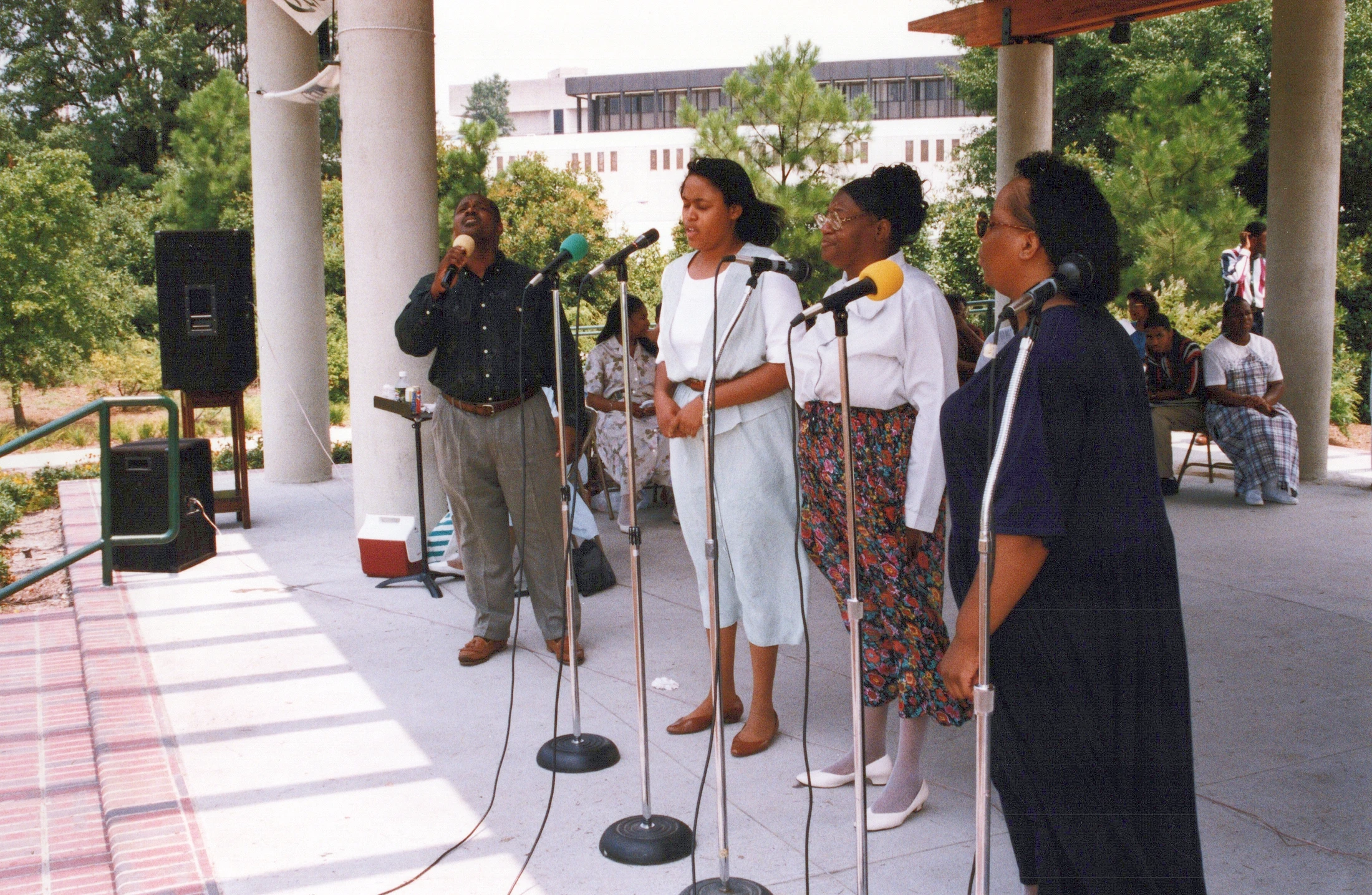 A group of four Black singers, one man and three women, stand in a line at microphones performing outdoors on a sunny day.
