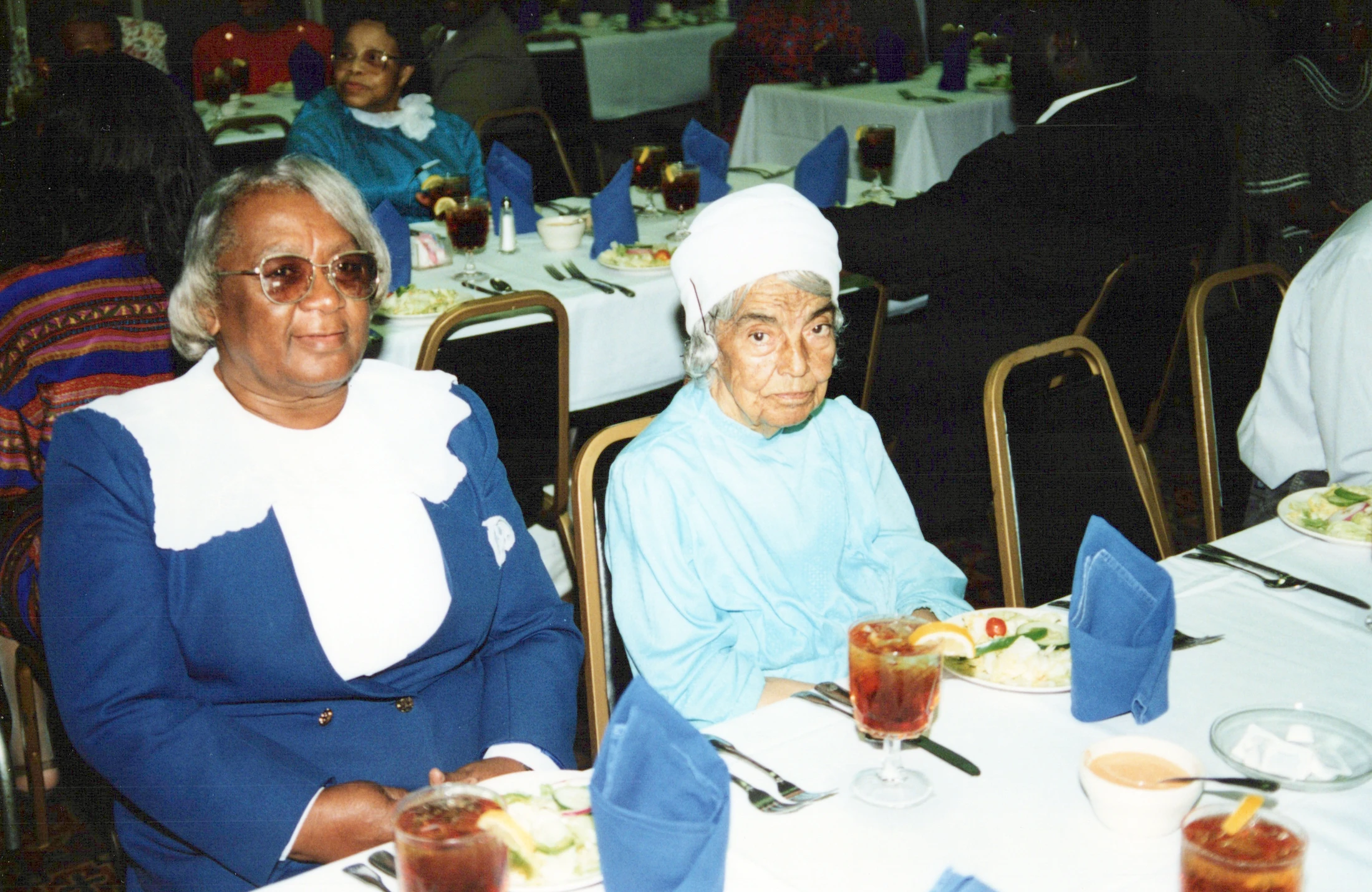 A vintage photograph of two older Black women seated at a dinner table during an event. The table is set with food, drinks, and blue cloth napkins.