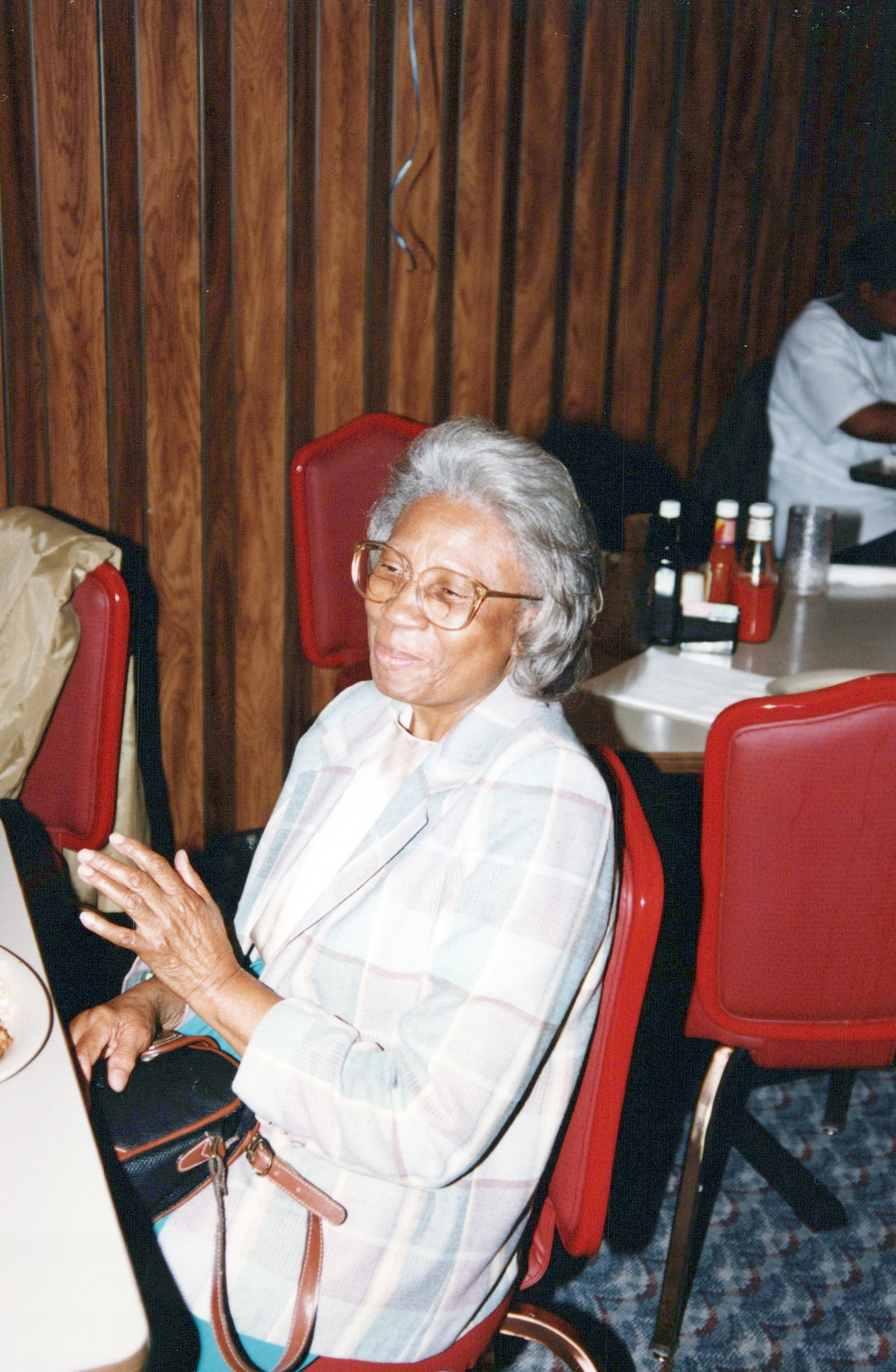 A vintage photograph of an elderly Black woman with gray hair and glasses, wearing a plaid jacket, sitting at a table in a restaurant with wood-paneled walls.
