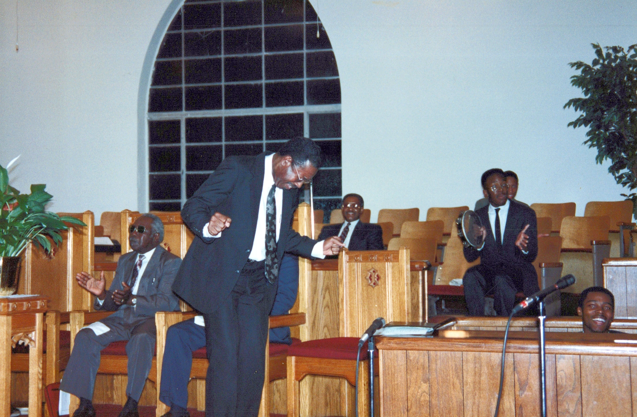 A Black man in a suit joyfully dances in a church aisle while other men in the congregation clap and play a tambourine from the pews.