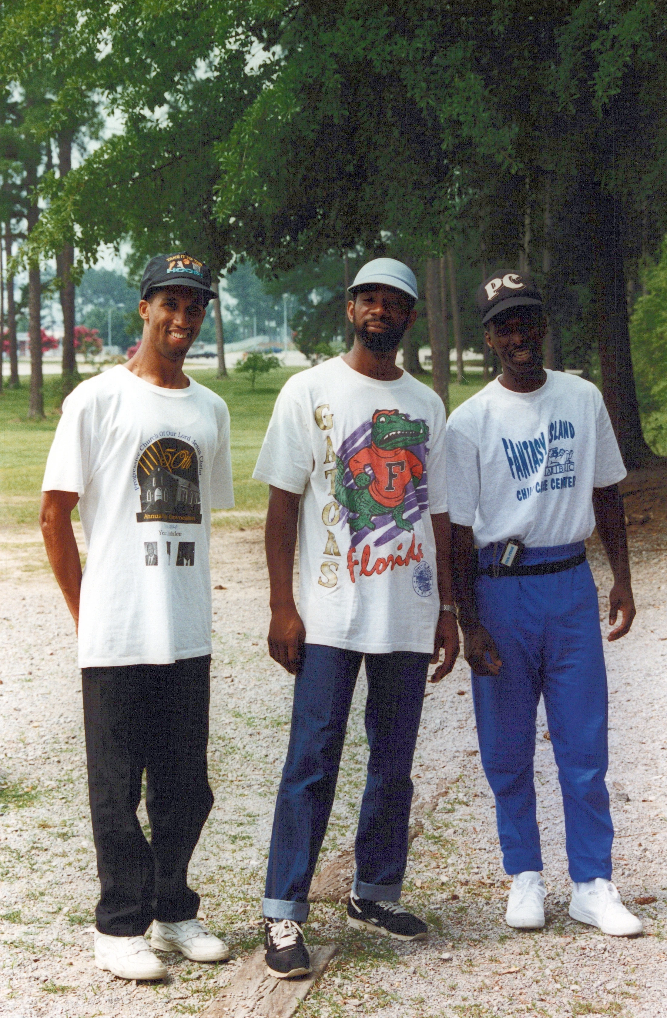 A vintage photograph of three African American men standing side-by-side in a park, wearing casual 1990s clothing like graphic t-shirts and baseball caps.