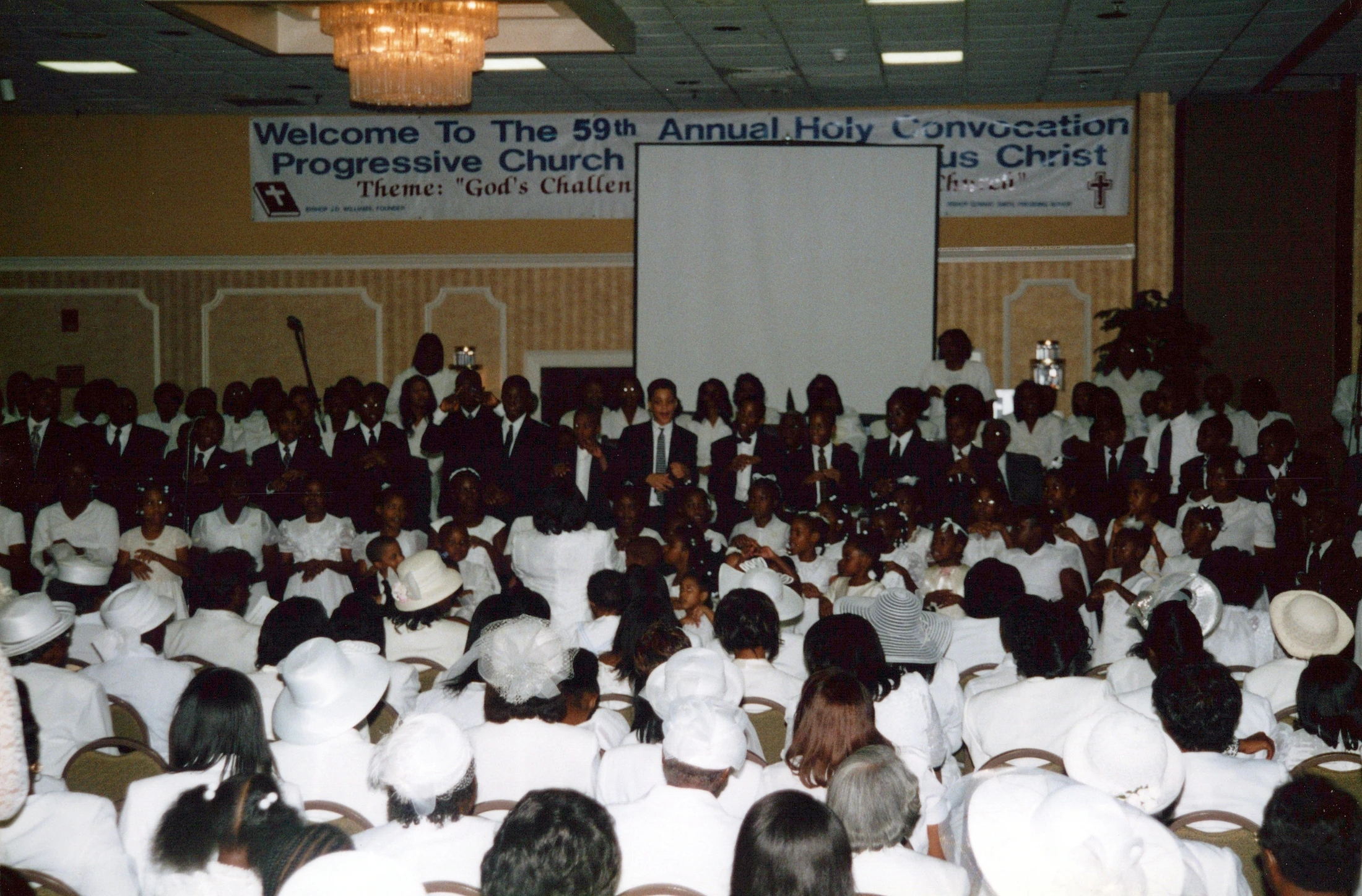 A large African American congregation, with many women in white, attend the 59th Annual Holy Convocation in a church hall with a banner in the background.