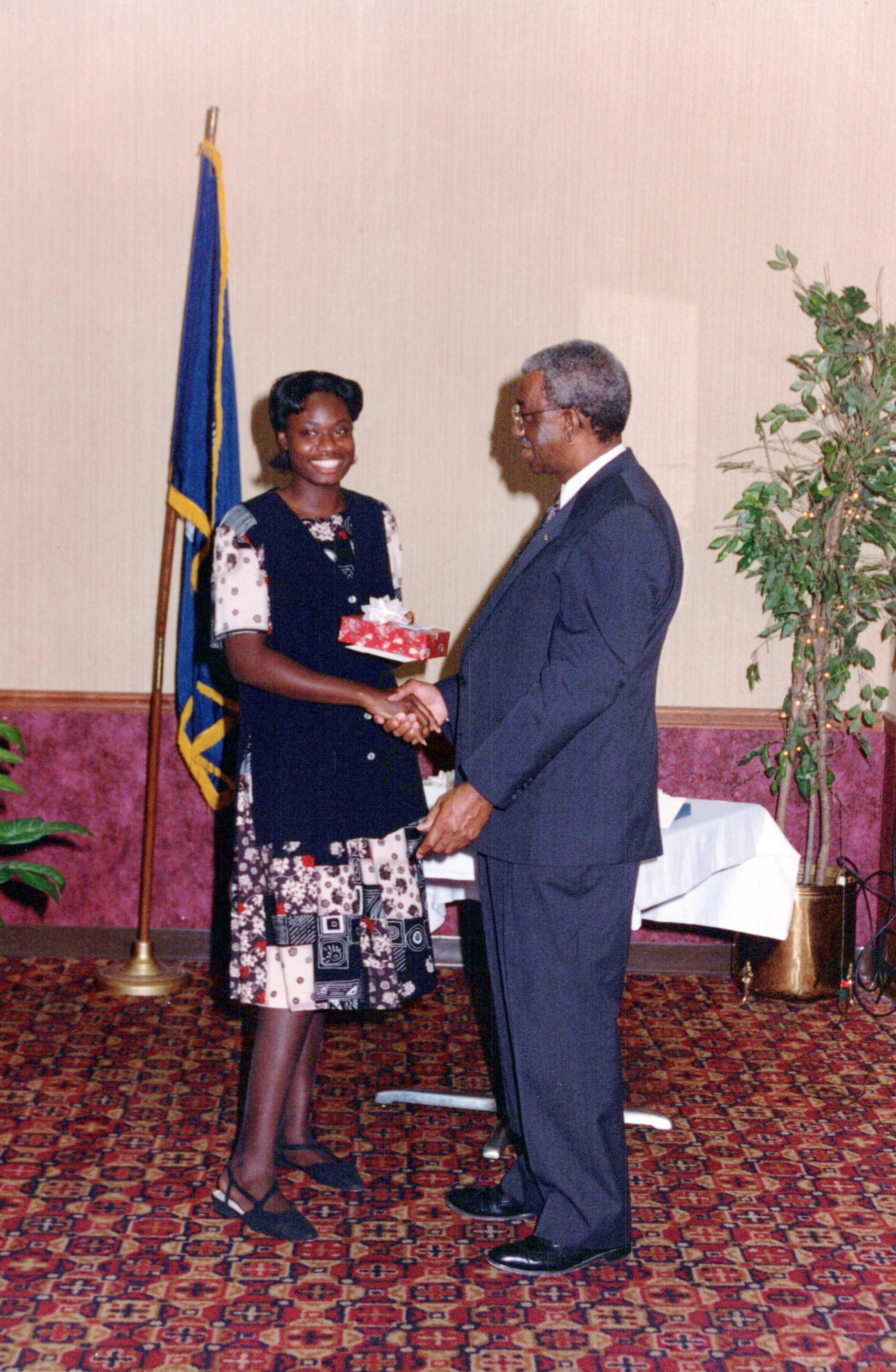 A smiling young Black woman in a patterned dress shakes hands with an older Black man in a suit. She holds a small, wrapped gift at a formal event.