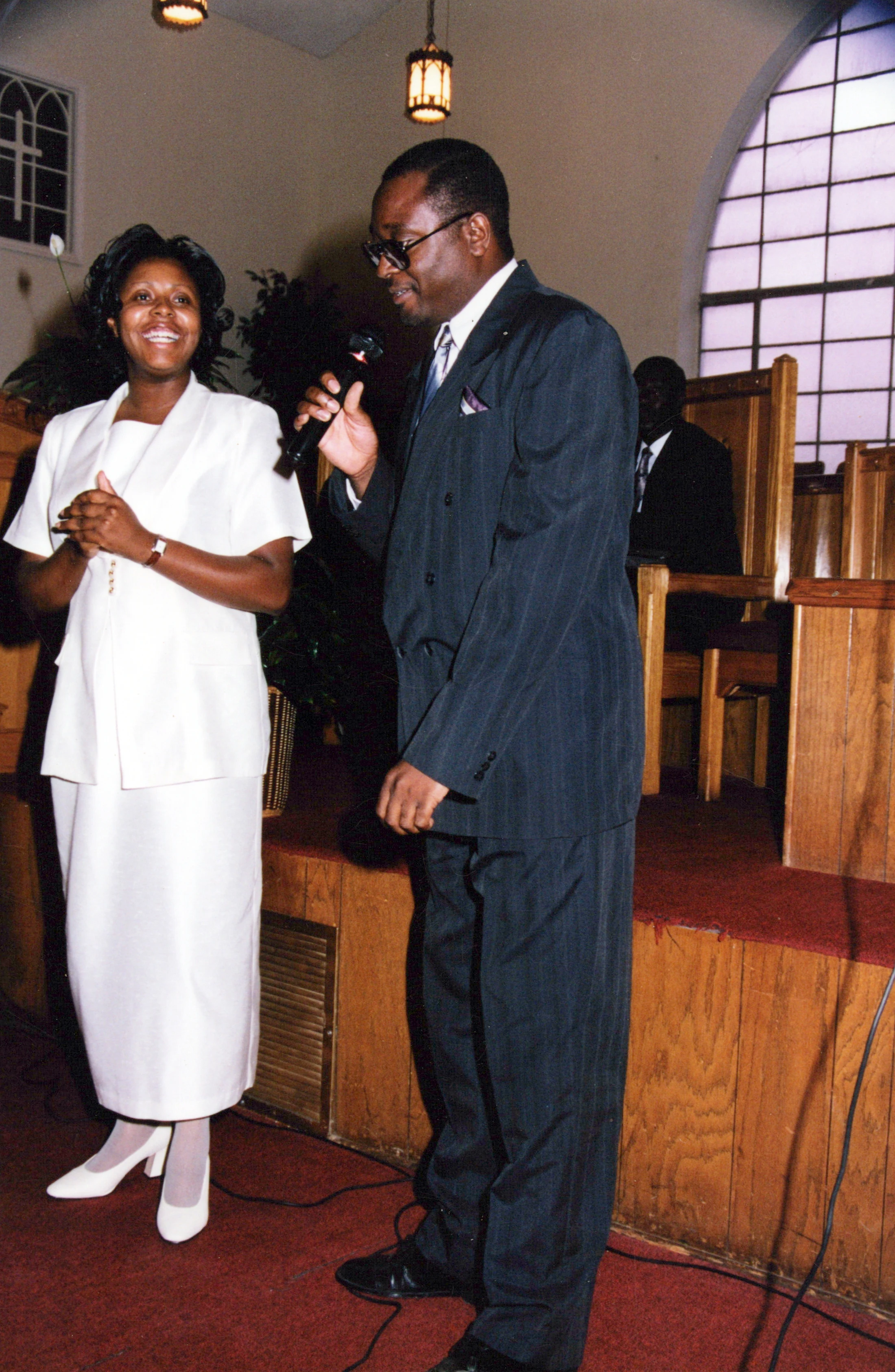 A man in a dark pinstripe suit speaks into a microphone next to a smiling woman in a white suit. They are standing on a red-carpeted stage in a church.