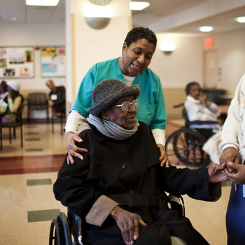 An elderly Black woman in a wheelchair is attended by a smiling nurse in scrubs and a nun holding her hand in a nursing home common area.