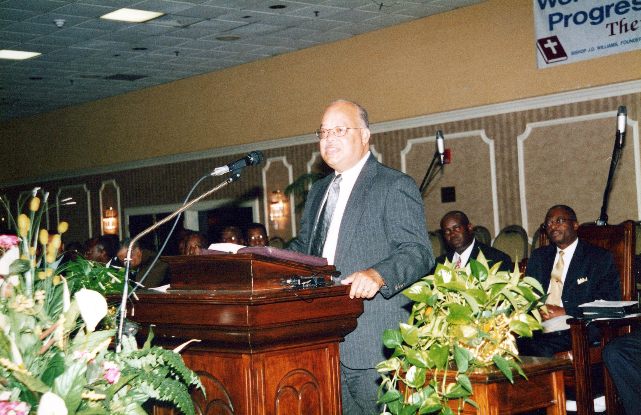 A smiling man with glasses, wearing a gray pinstripe suit, stands at a wooden lectern and speaks into a microphone. Two men are seated behind him.