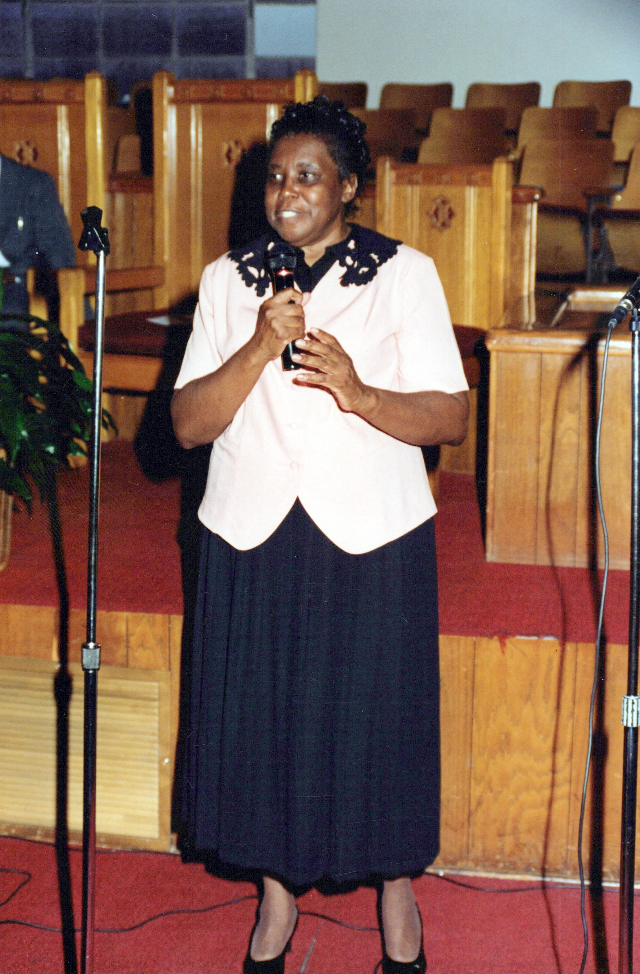 A smiling Black woman in a pink shirt and black skirt stands on a red-carpeted stage in a church, holding a microphone and speaking.