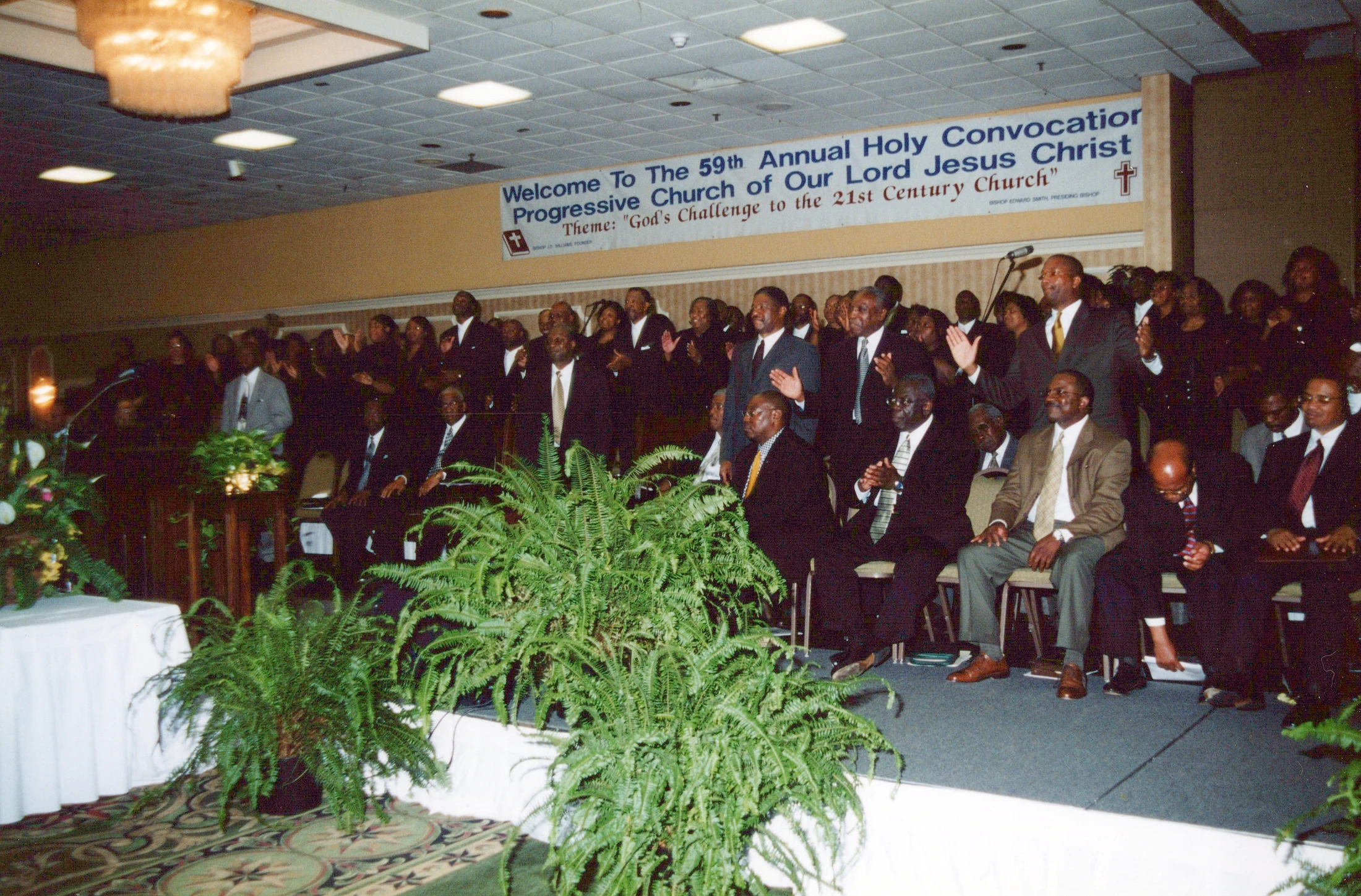 A large congregation of mostly African American men in suits at a church convocation. Many are standing and singing while others are seated on a stage.