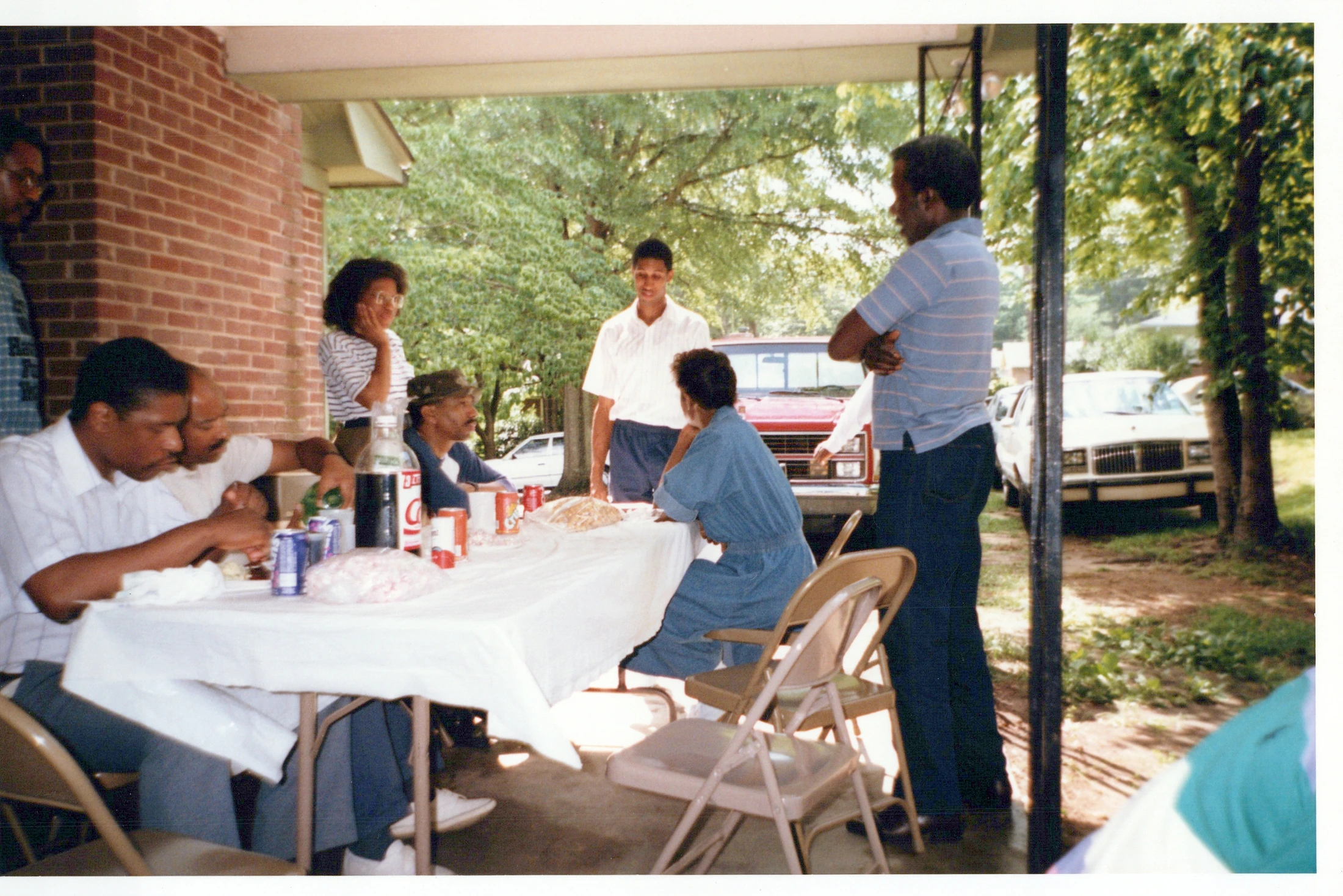 A vintage color photograph of a Black family gathering for a meal outdoors. People sit and stand around a long table with food on a covered patio.