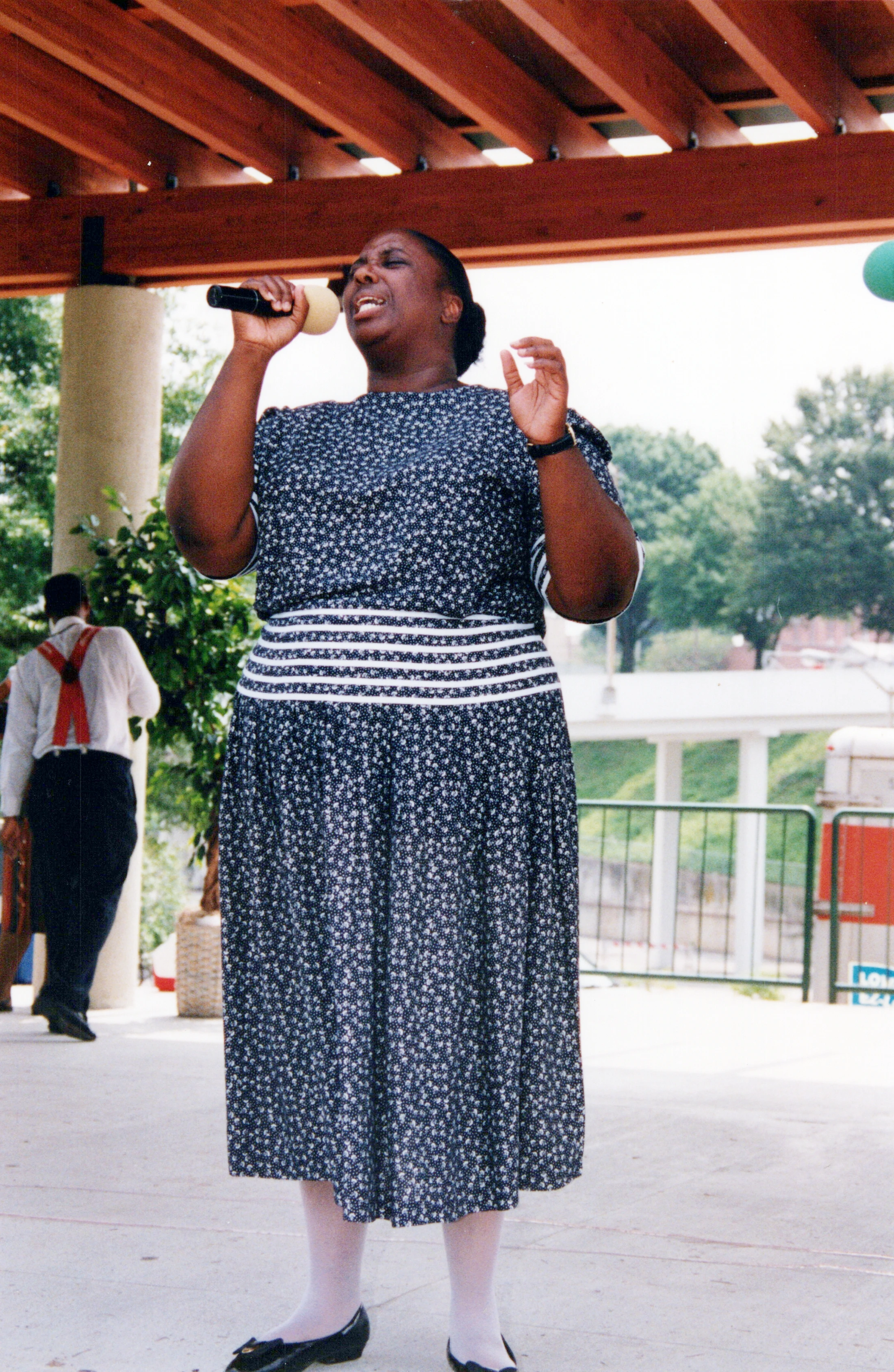 An African American woman in a patterned dress sings passionately into a microphone while standing outdoors under a wooden structure.