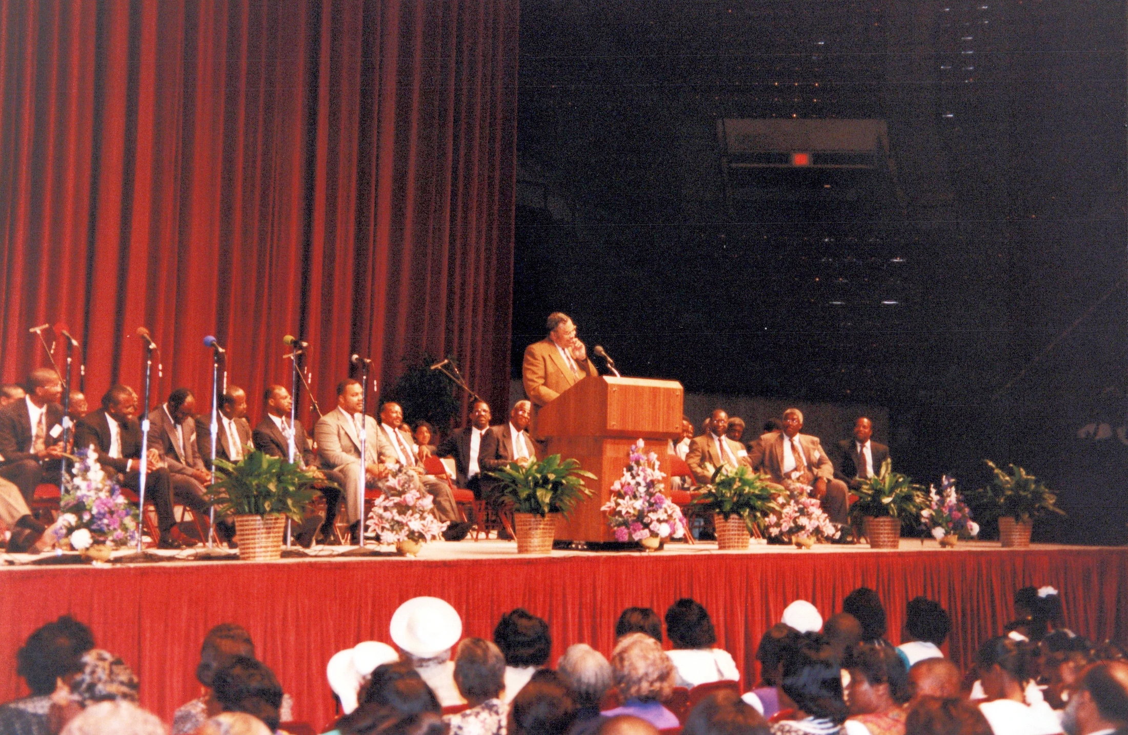 A man in a suit speaks at a wooden podium on a stage in front of a red curtain. Other men sit behind him on stage, and an audience watches from below.