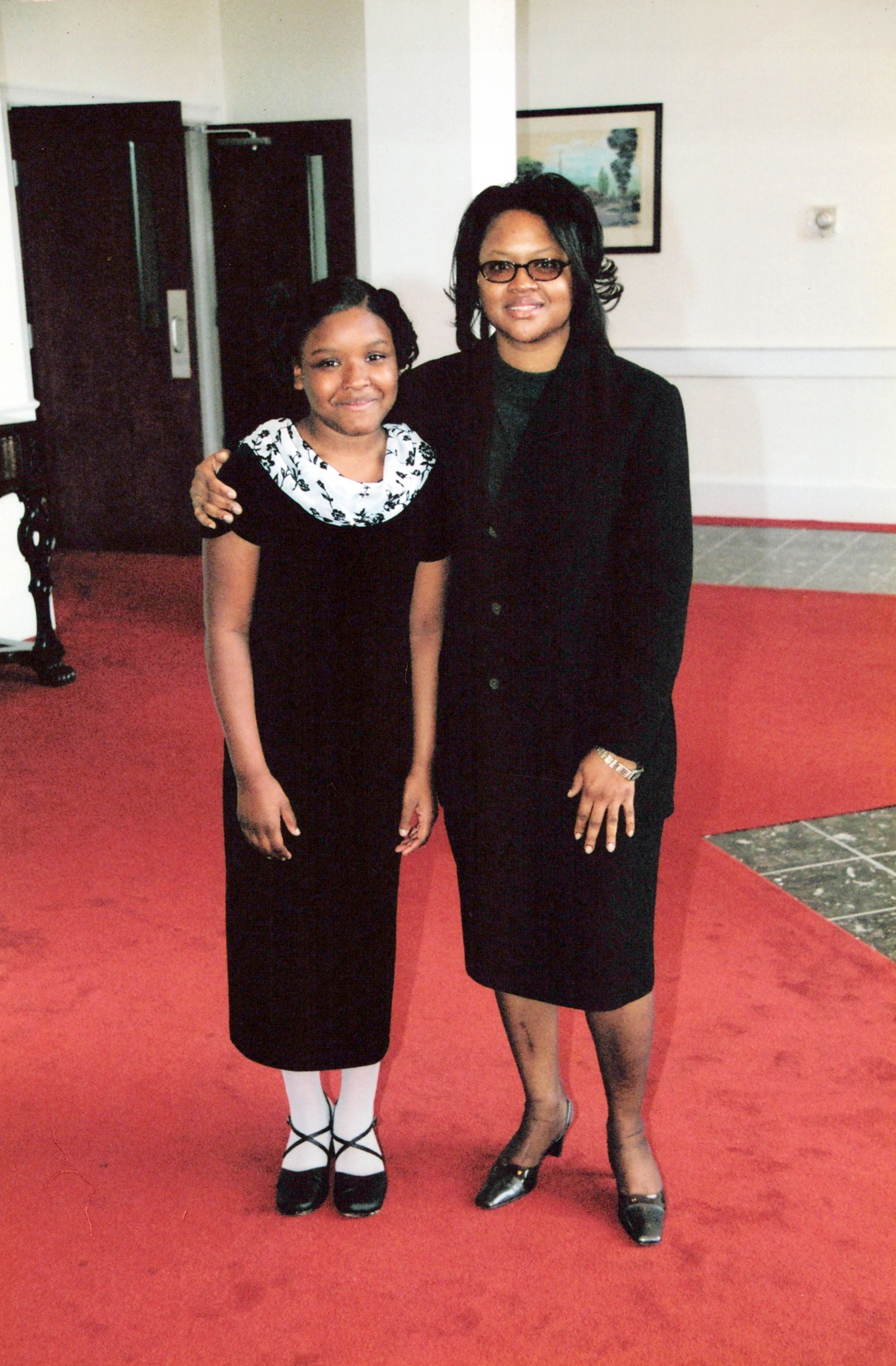 A Black woman and a young Black girl stand smiling for a photo on a red carpet. Both are wearing black formal outfits. The woman has her arm around the girl.