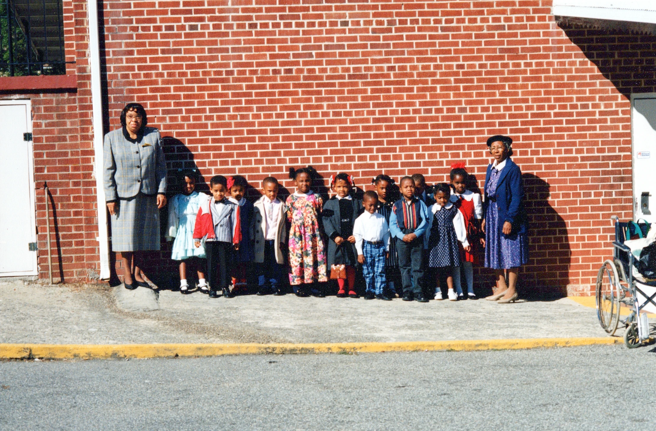 A group of young African American children and two female teachers in suits pose for a photo outdoors, standing in a line against a red brick wall.