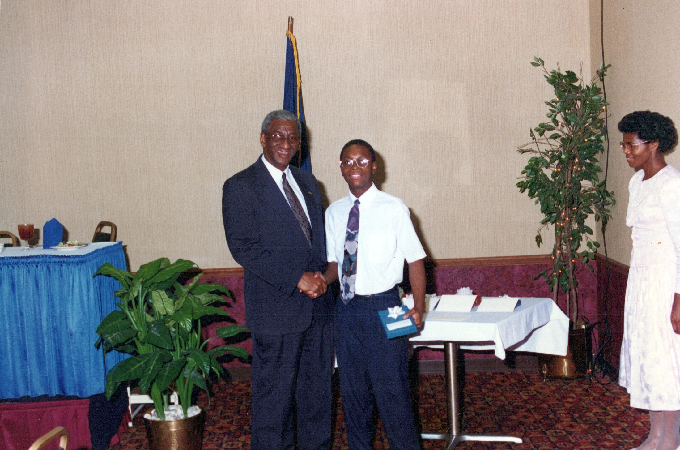 An older man in a suit shakes hands with a smiling young man in a shirt and tie. The young man holds a small gift box. A woman looks on from the side.