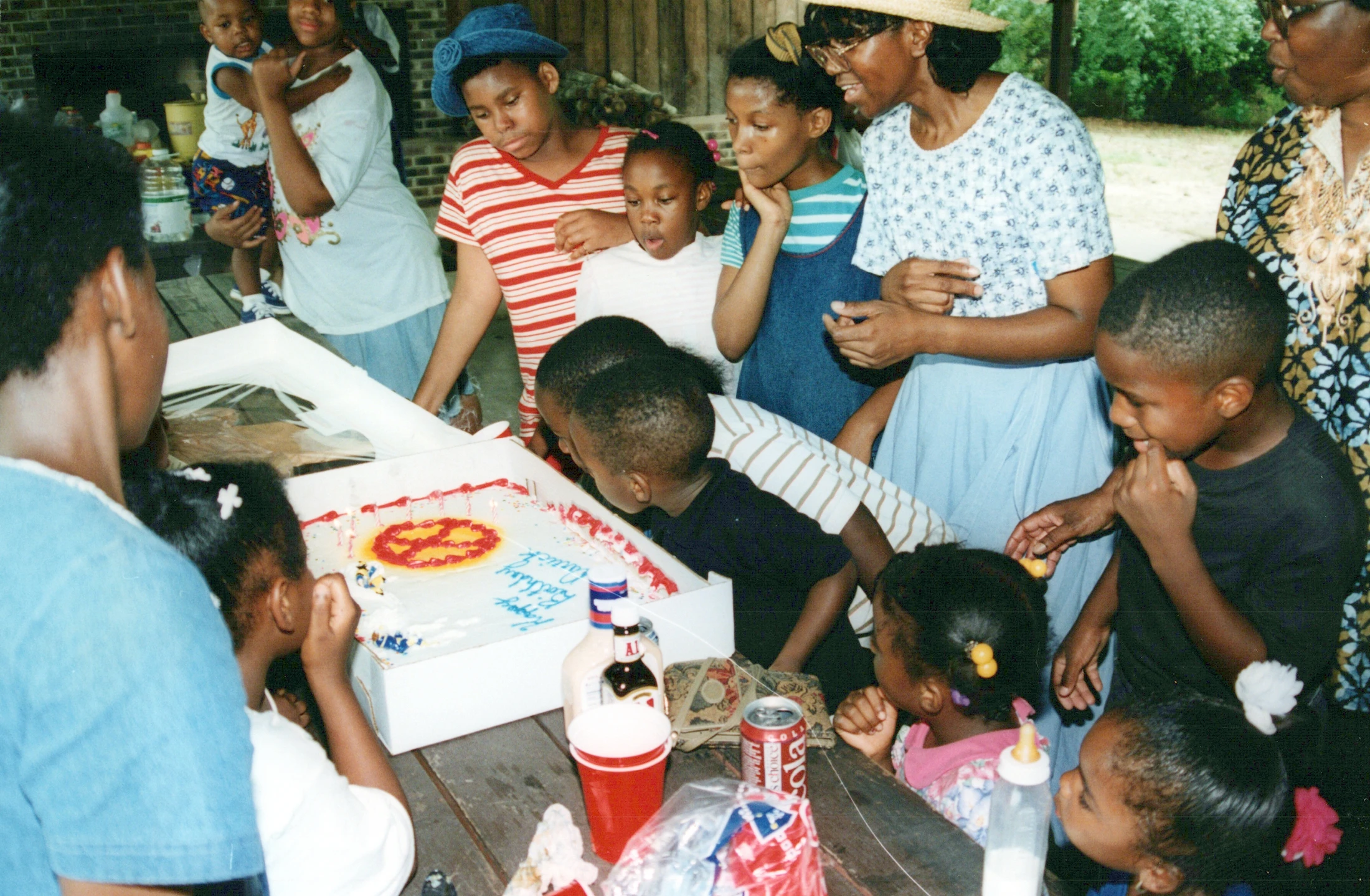 A large Black family with adults and children gathers around a wooden picnic table, looking at a white birthday cake with red and yellow frosting.