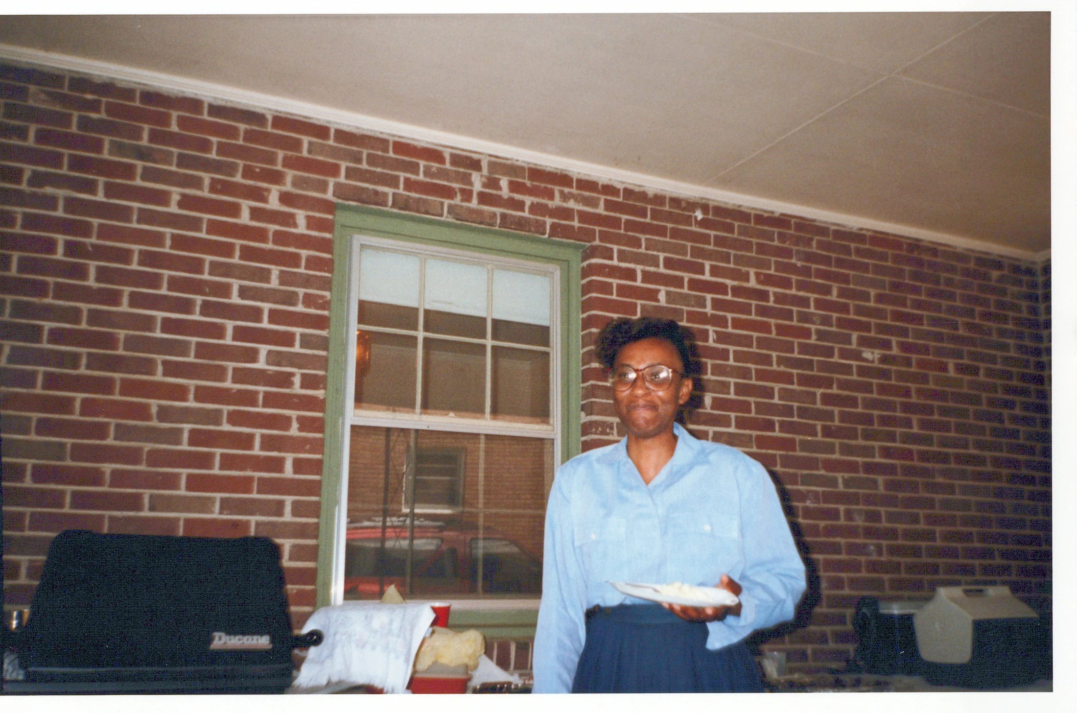 A vintage photograph of a Black woman with glasses, smiling and holding a plate of food while standing on a brick patio in front of a house.