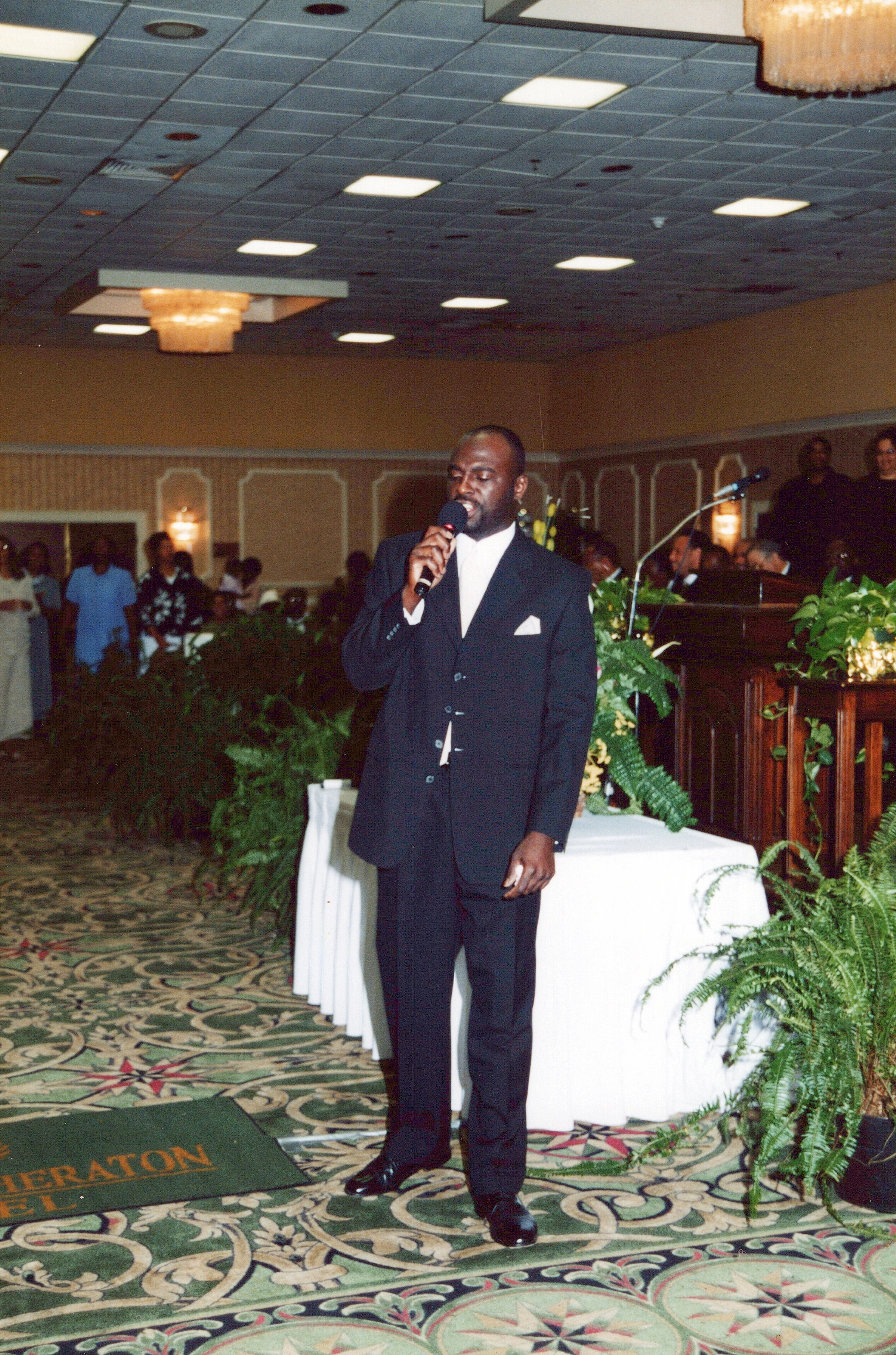 A Black man in a dark suit and white shirt stands on a patterned carpet in a hotel ballroom, speaking into a microphone.